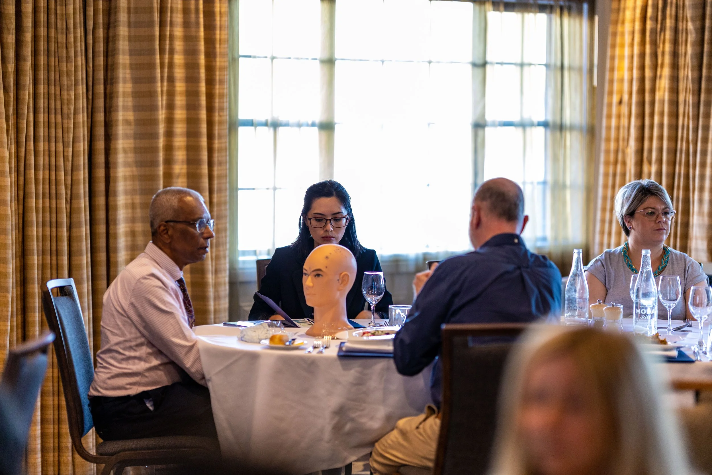 A group of four people sitting at a round table in a well-lit room with large windows and yellow curtains, engaged in a discussion or activity, with a mannequin head on the table.