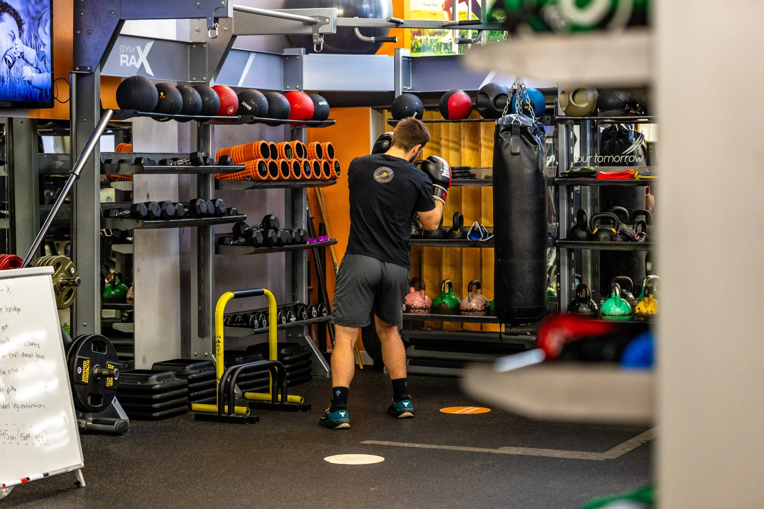 A man in a black T-shirt and gray shorts boxers at a boxing gym, practicing with black boxing gloves. The gym has various weights, kettlebells, medicine balls, and punching bags on shelves and around the room.