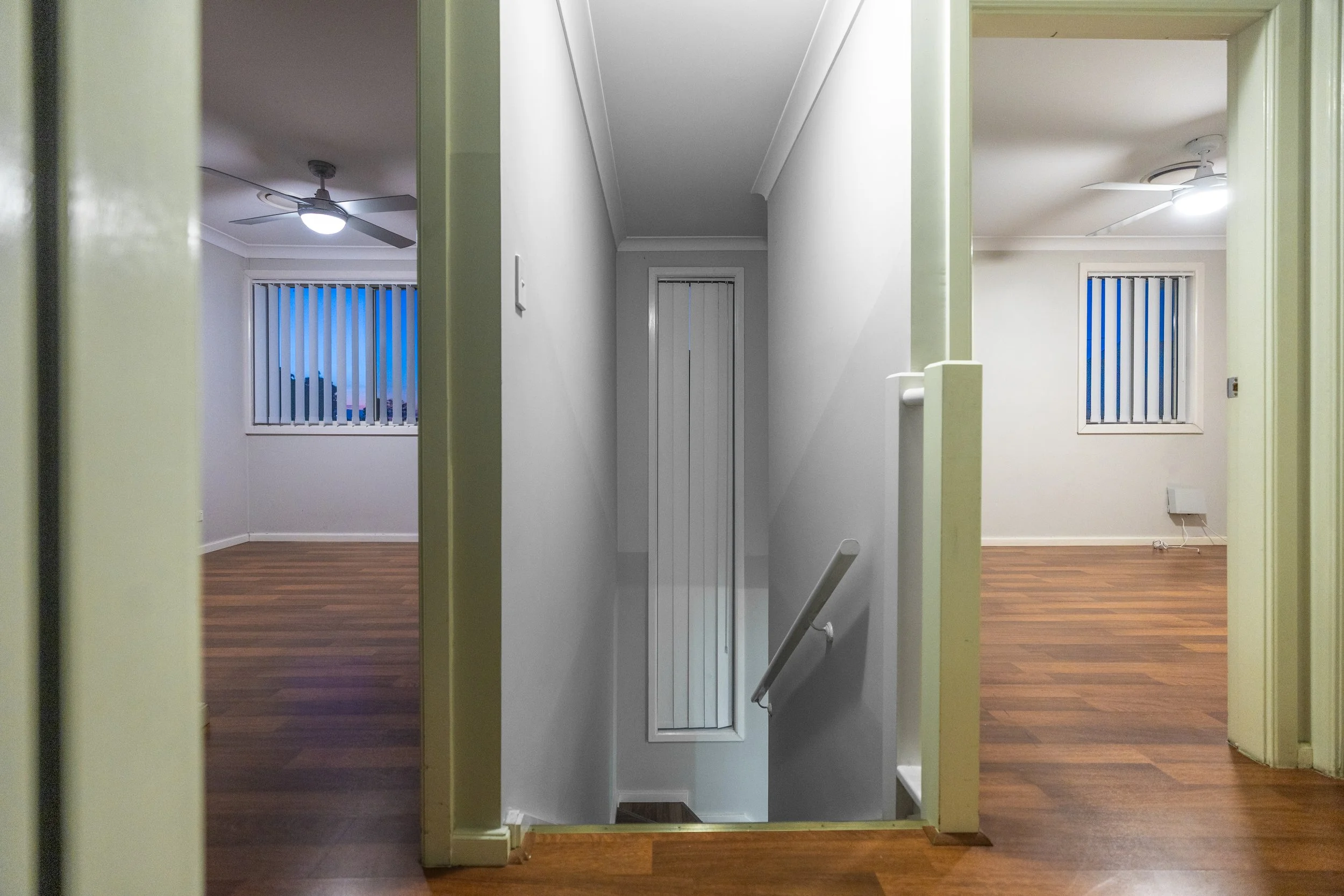 Interior view of a stairwell with brown hardwood floors, white walls, and a ceiling with ceiling fans and lights; two rooms with windows and vertical blinds are visible on either side.
