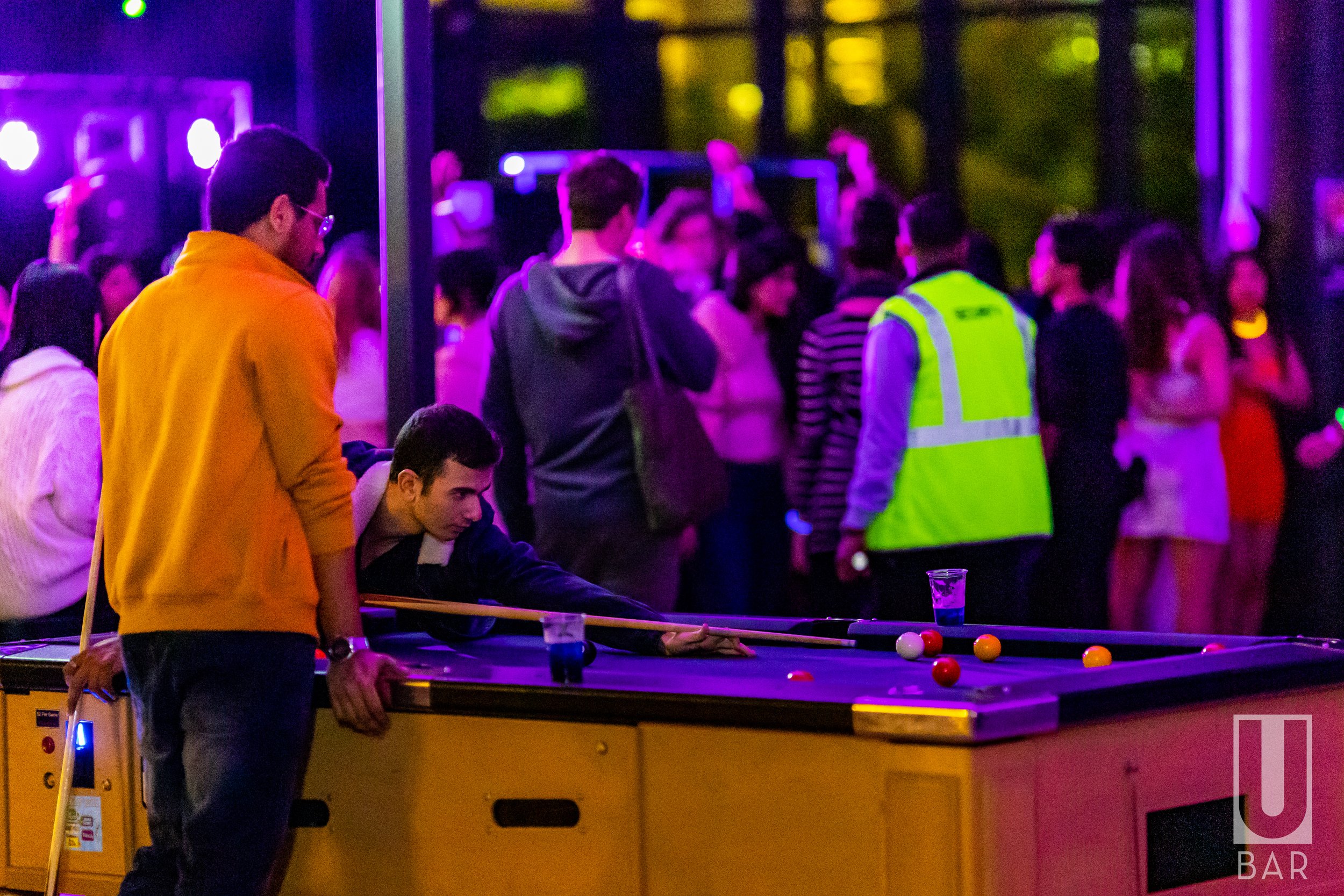 People playing pool at a bar with vibrant purple and yellow lighting, in a crowded social setting.
