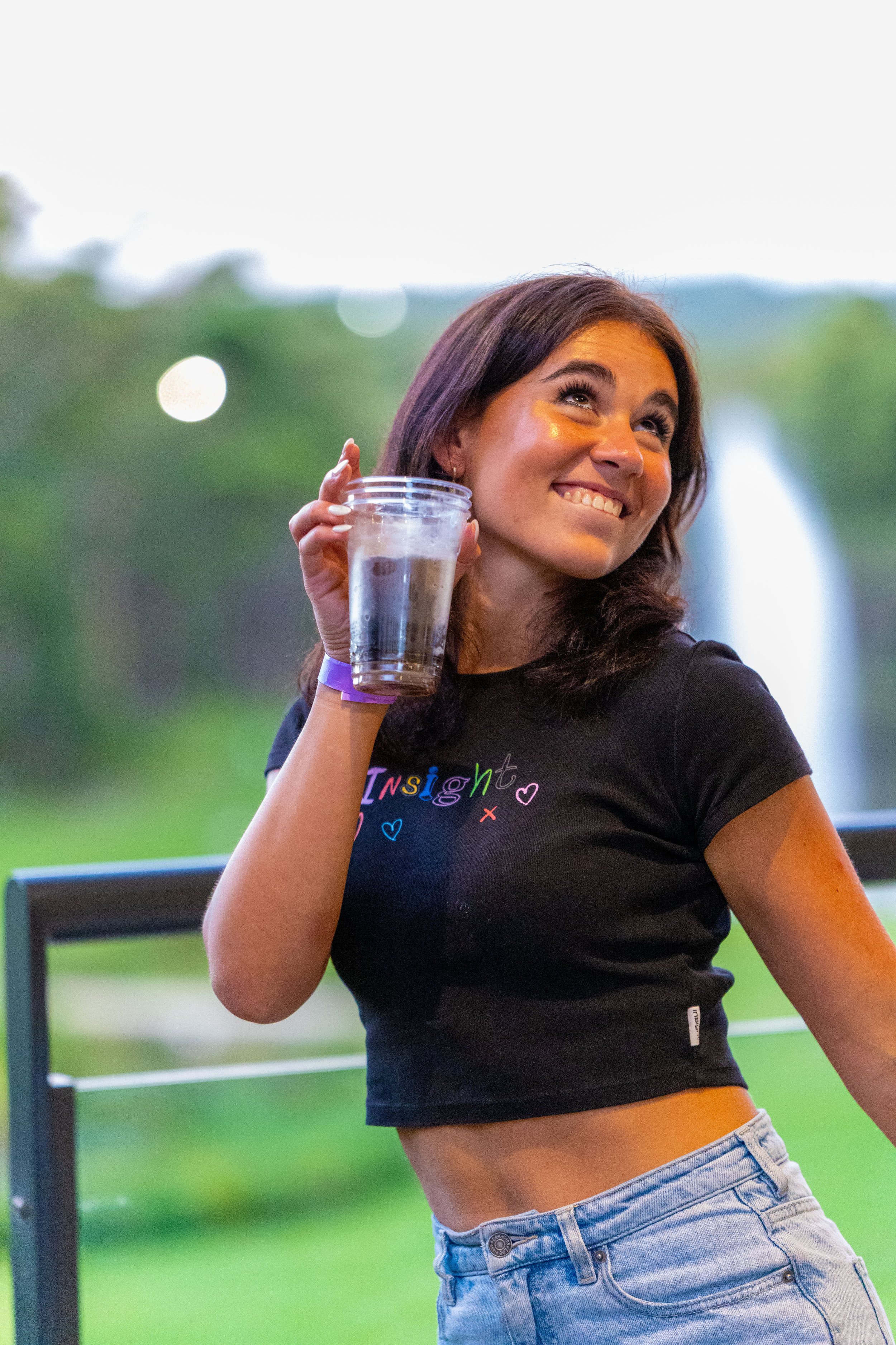 A young woman with dark hair and a bright smile holds a clear plastic cup with water, standing outdoors with a blurred green background and a water fountain in the distance.