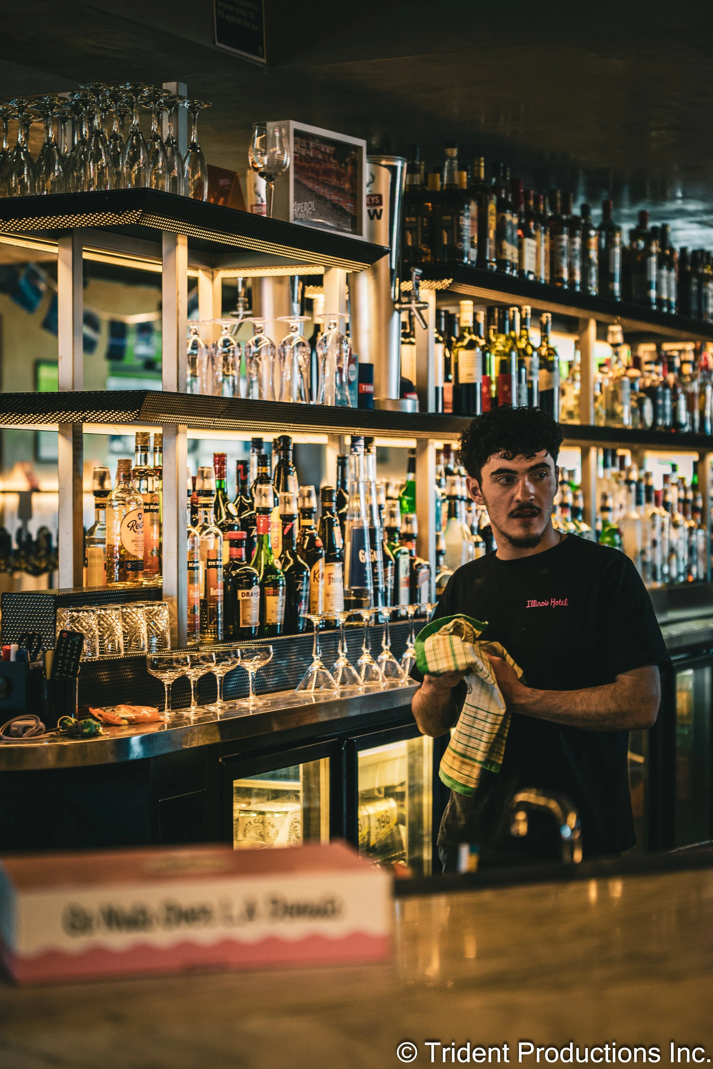 A bartender with curly hair and a black t-shirt wiping a glass with a cloth behind a bar counter stocked with various bottles of alcohol and glassware.