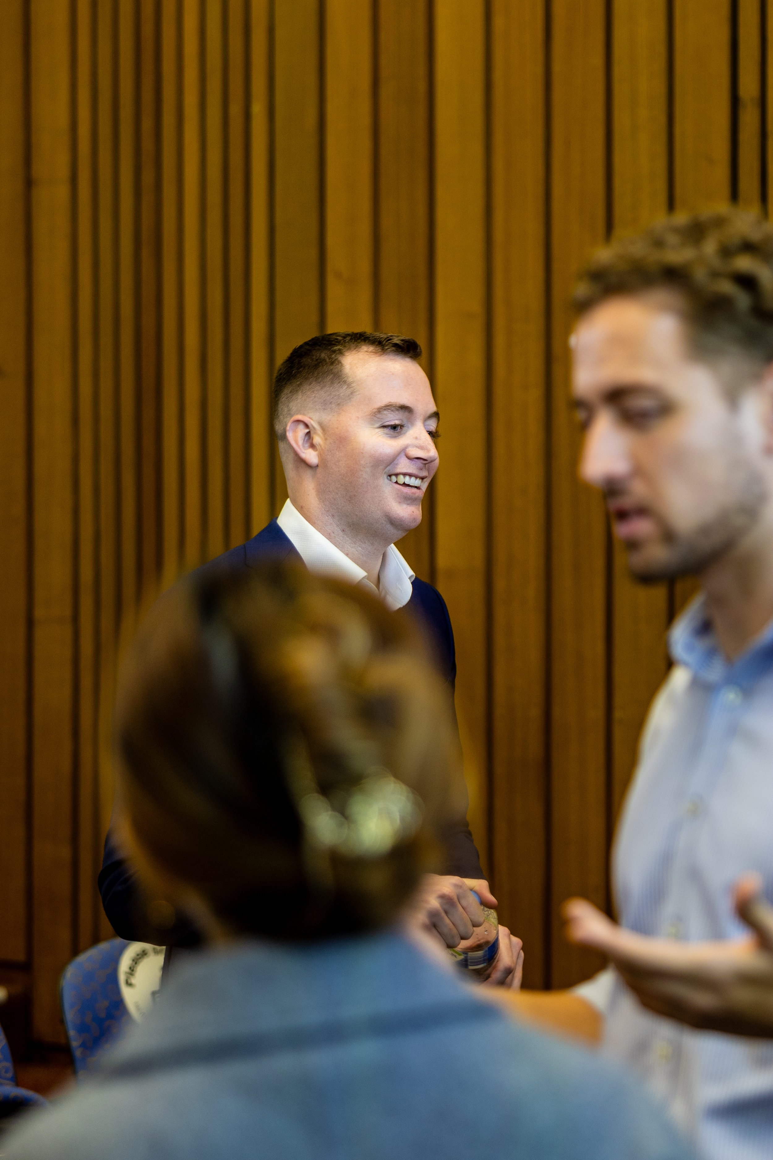 A young man in a dark suit smiling and talking to others at a social event with wooden paneling in the background.