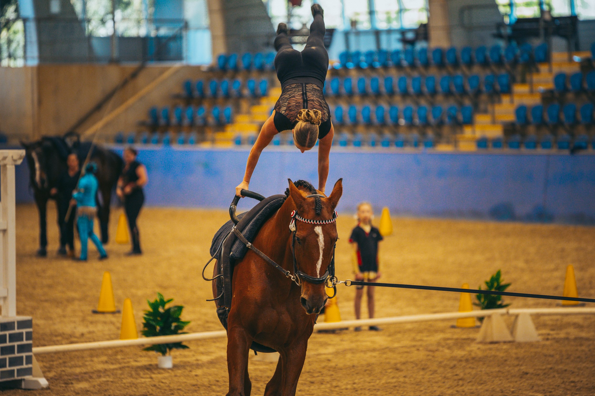 A female gymnast performing a handstand on a horse in an indoor arena with seats and cones in the background.