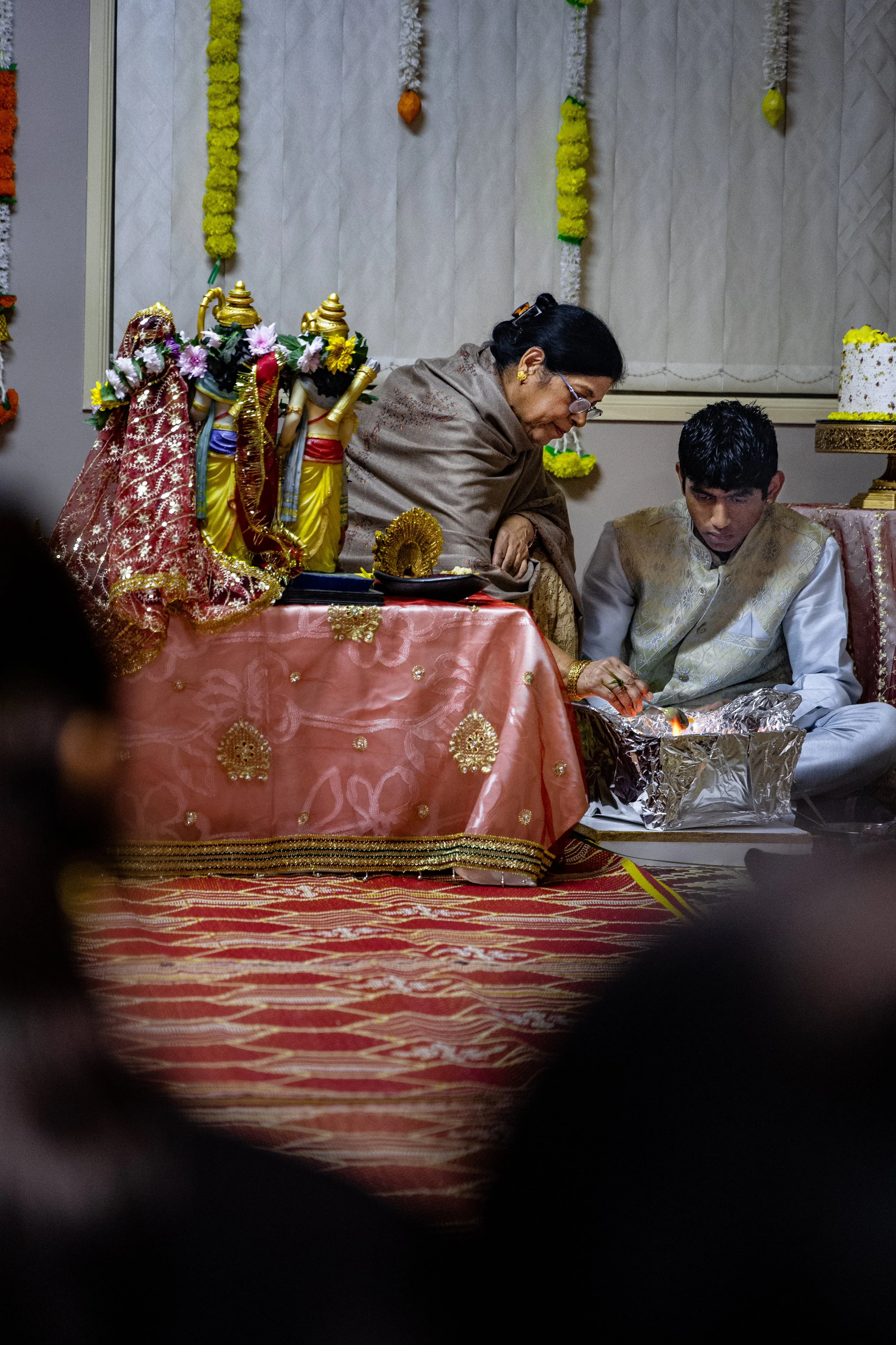 A traditional Indian wedding ceremony featuring a woman and a young man sitting on a patterned red carpet, with religious idols decorated with flowers on a pink tablecloth nearby. The woman appears to be performing a ritual, with the young man partic