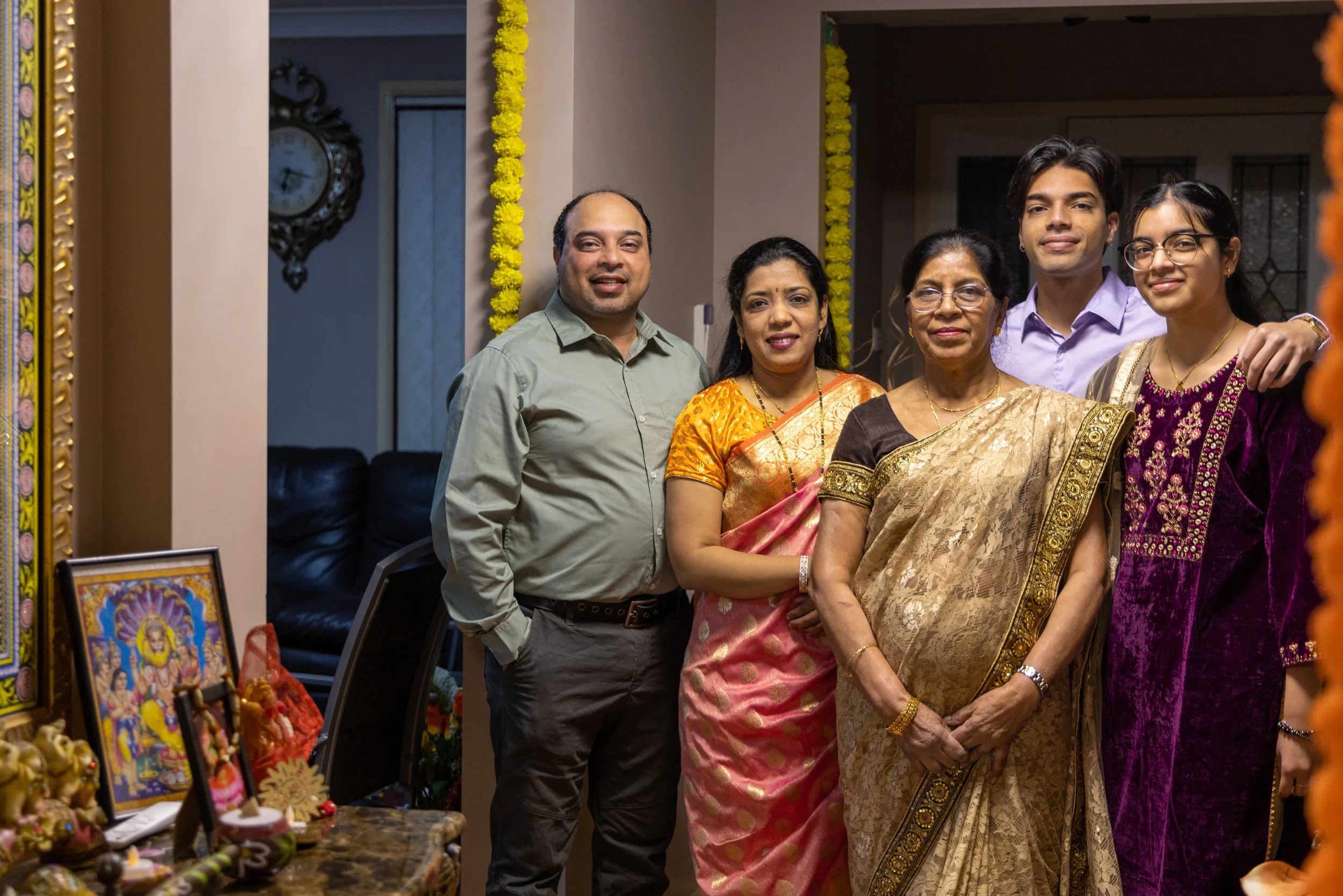A group of six people, including both men and women, dressed in traditional Indian attire, standing together indoors at a celebration, with decorations and religious images on a table nearby.