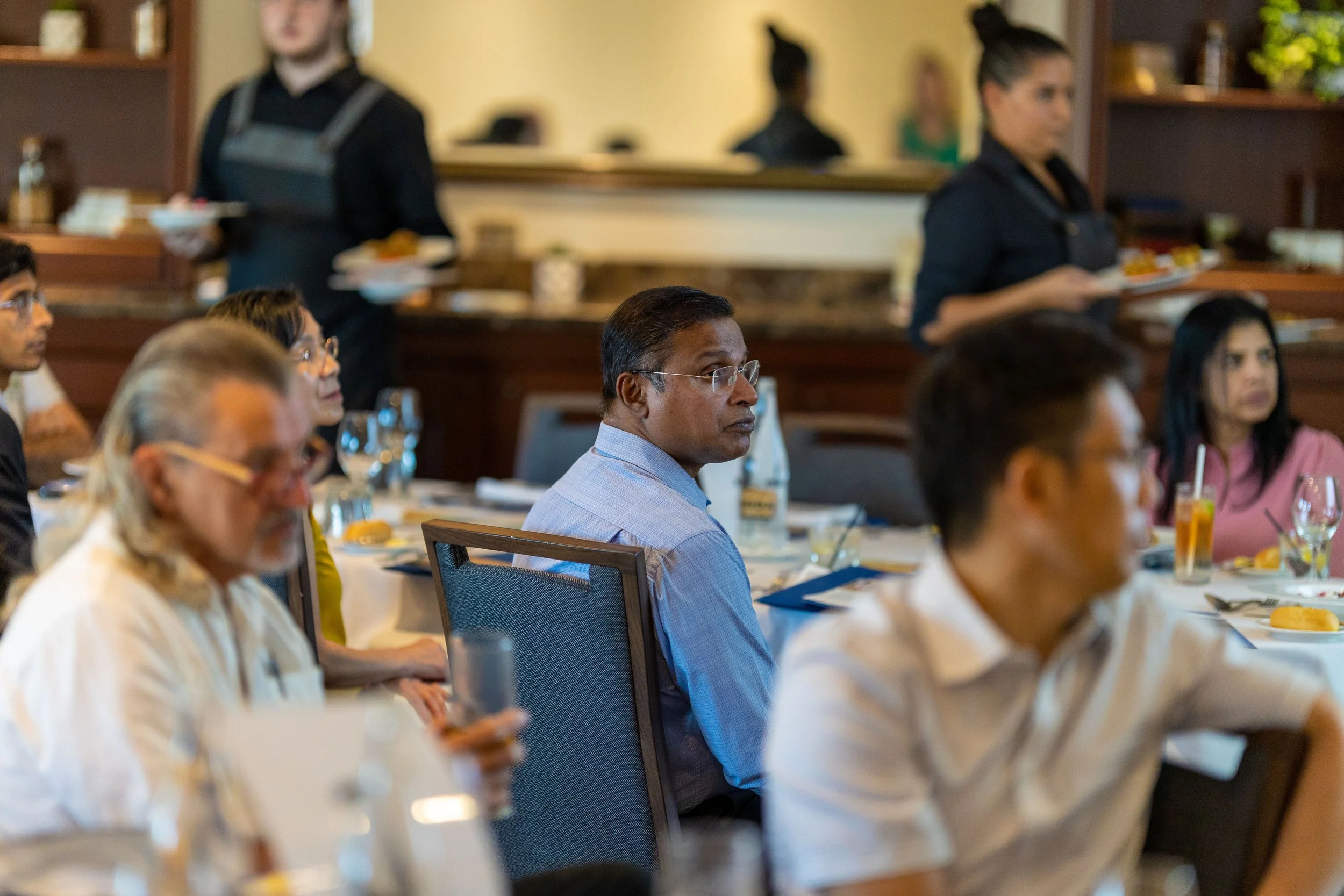 People attending a formal meal or conference, seated at tables and listening attentively, with waitstaff serving food in the background.