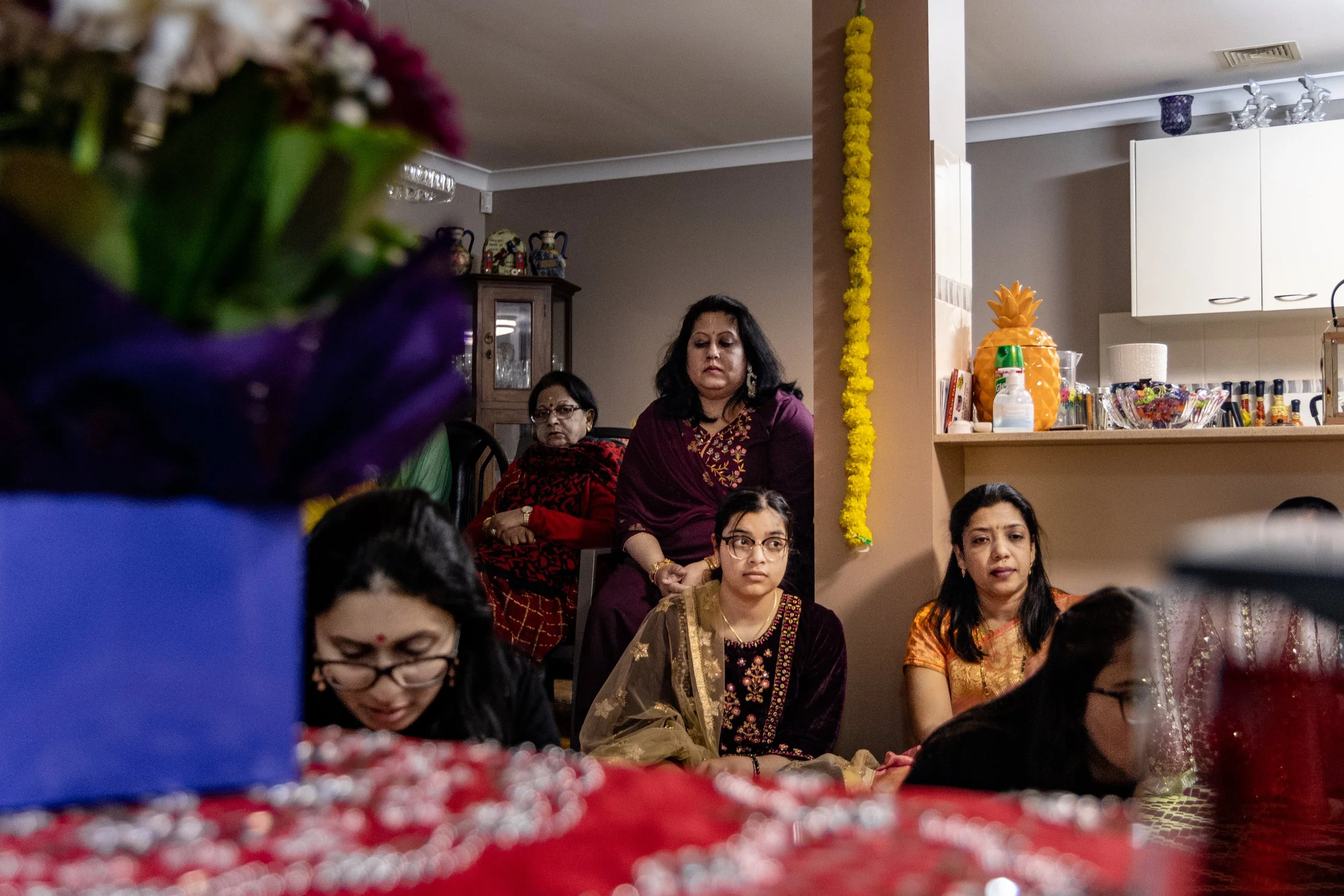 Group of women dressed in traditional Indian attire attending a cultural or religious gathering in a home setting, with decorative flowers and food items visible.