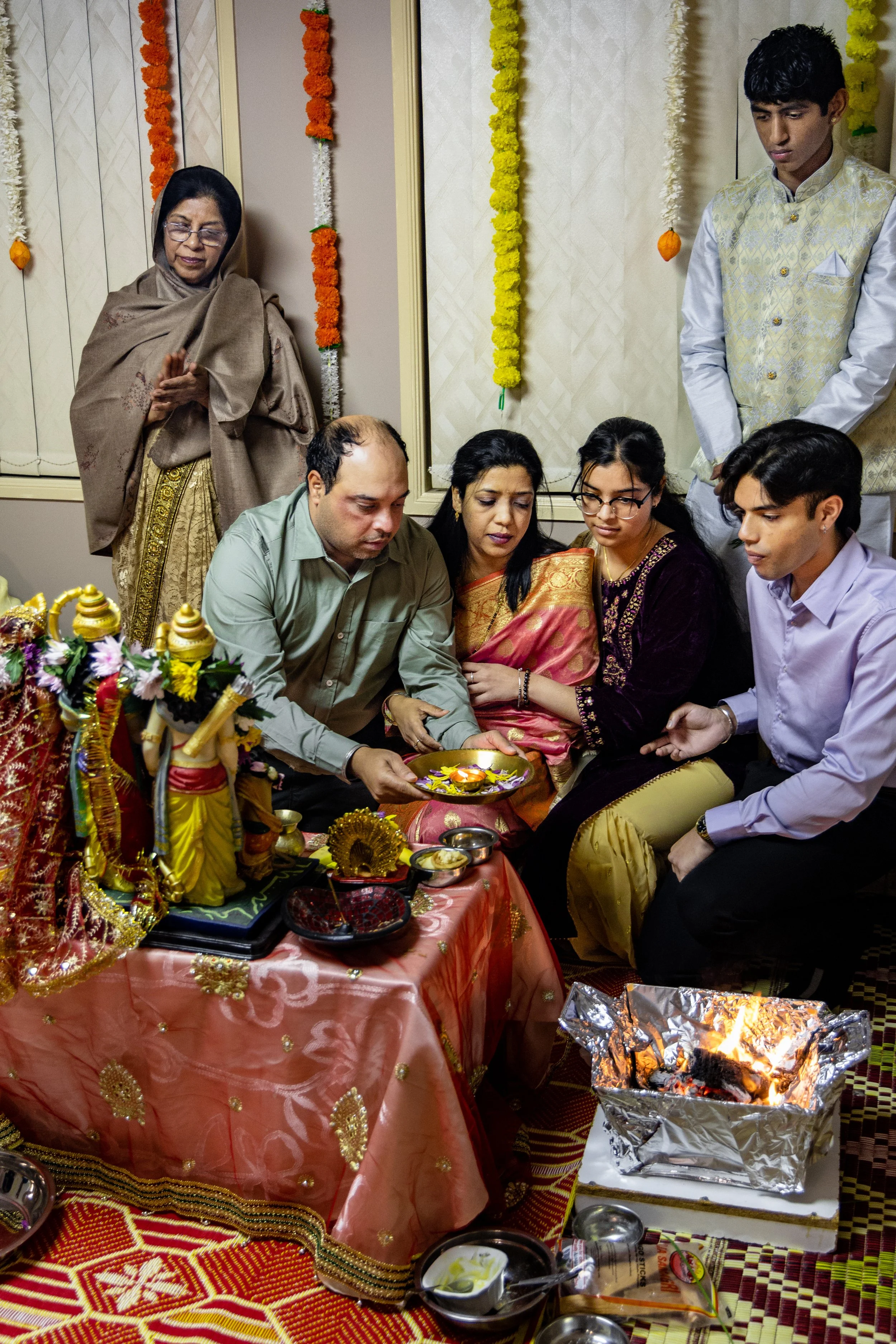 People performing a traditional Hindu puja ceremony, with offerings and a fire ritual, decorated with garlands and religious idols in the background.