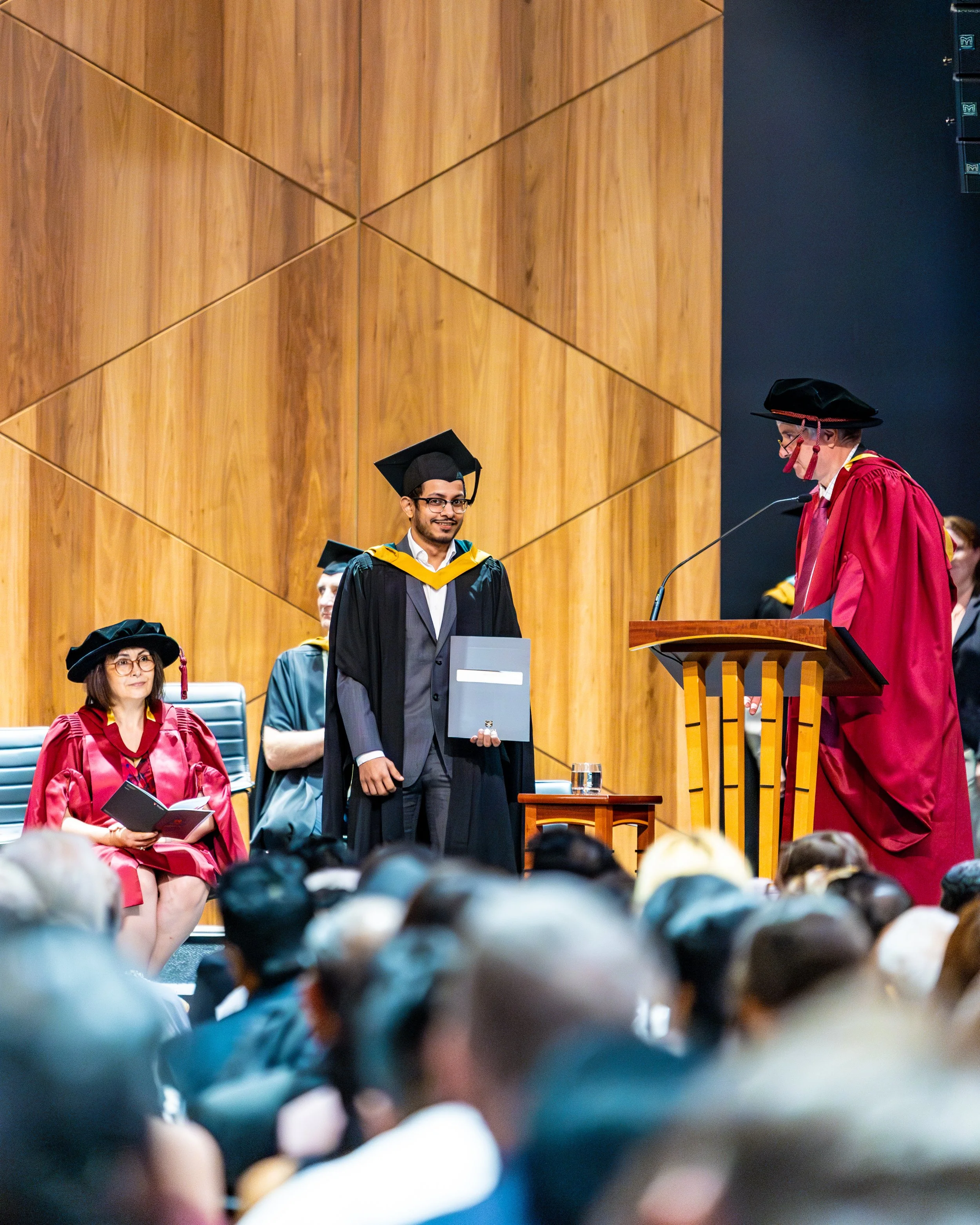 A graduation ceremony with a graduate in a tuxedo and cap receiving his diploma from a faculty member in a red gown and black hat on stage. Others in academic regalia are seated and the audience watches.
