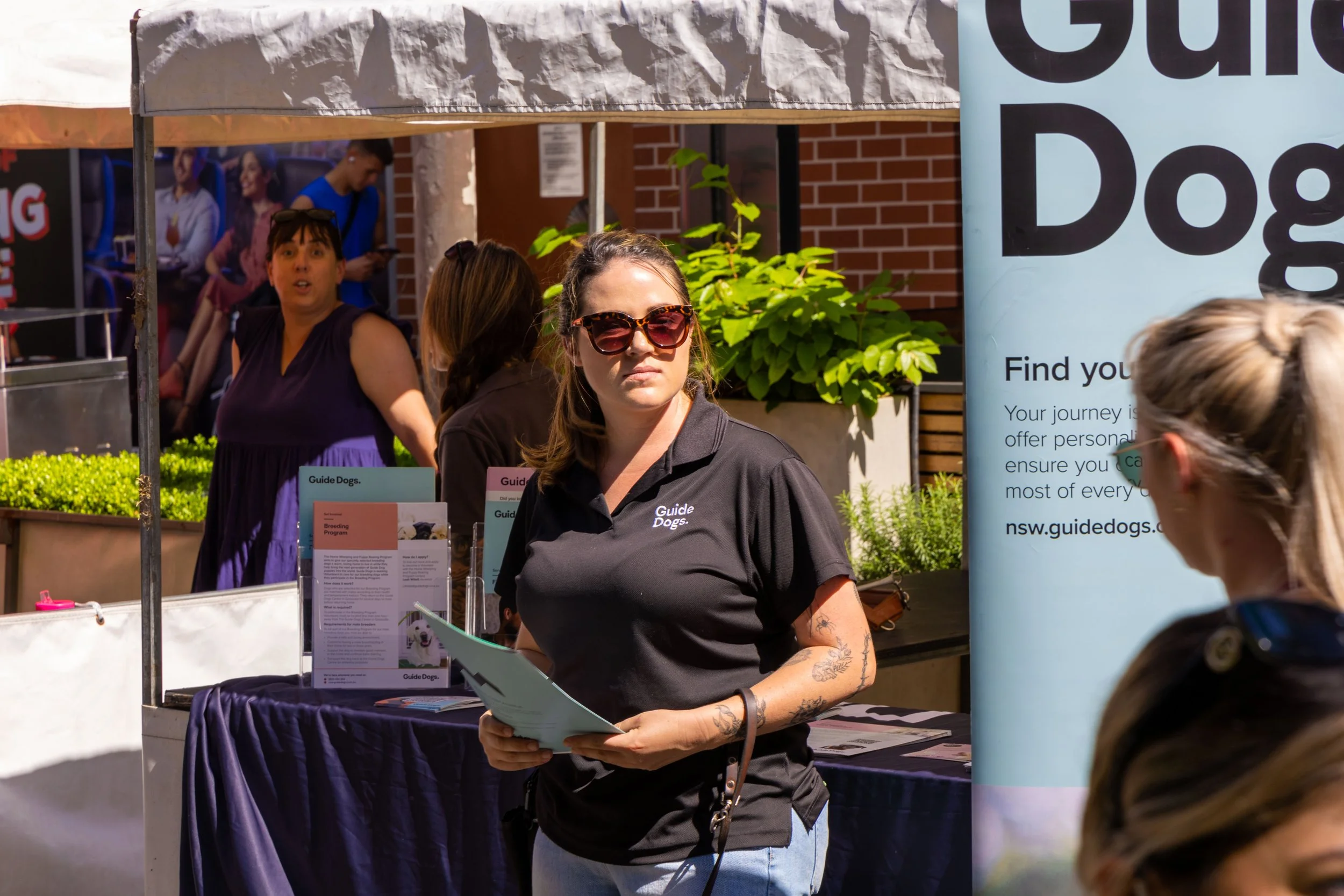 A woman in a black shirt with sunglasses holding informational brochures at an outdoor booth for Guide Dogs, with people and a banner in the background.