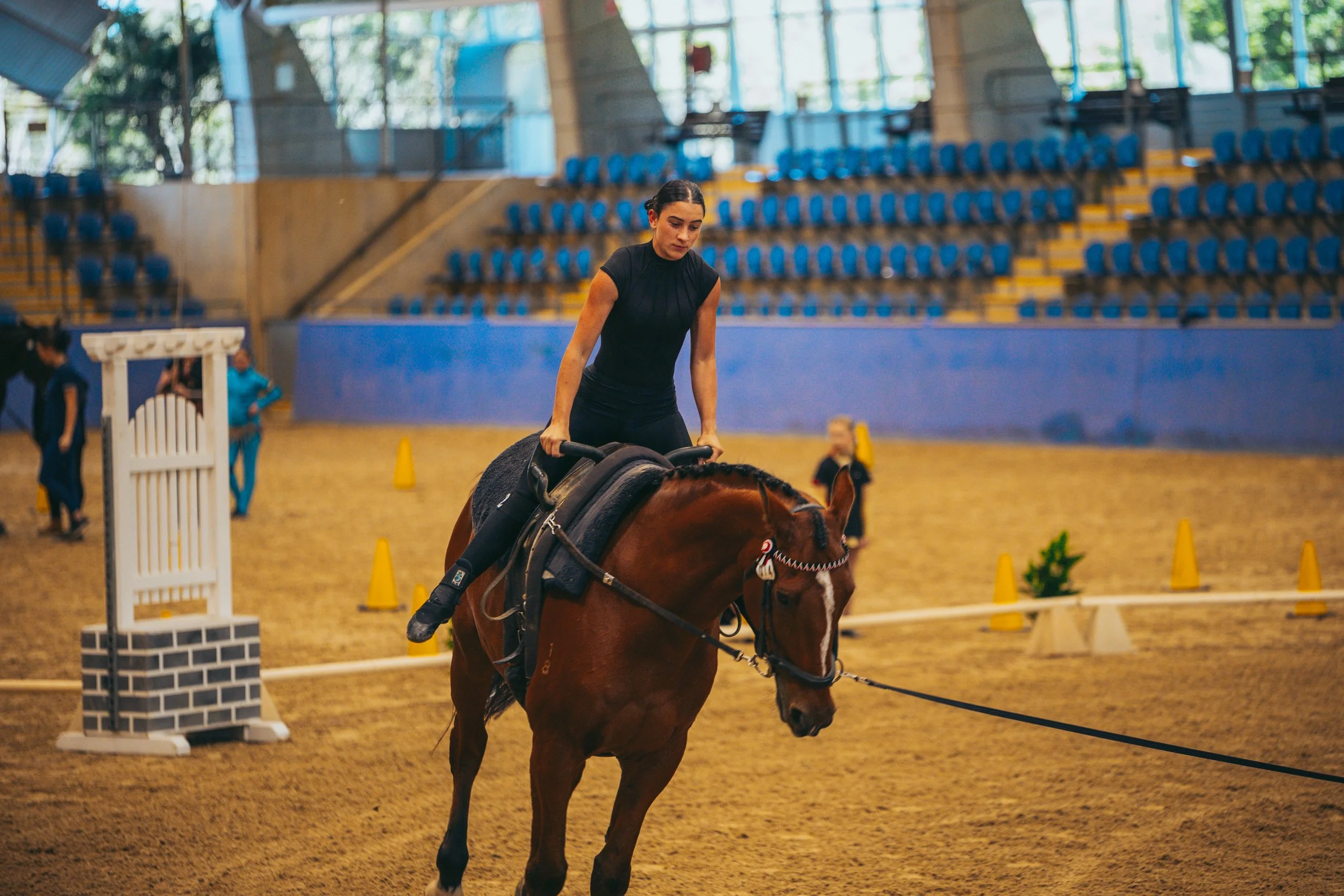 A woman riding a horse inside an indoorized equestrian arena, practicing or training, with empty blue seats in the background.