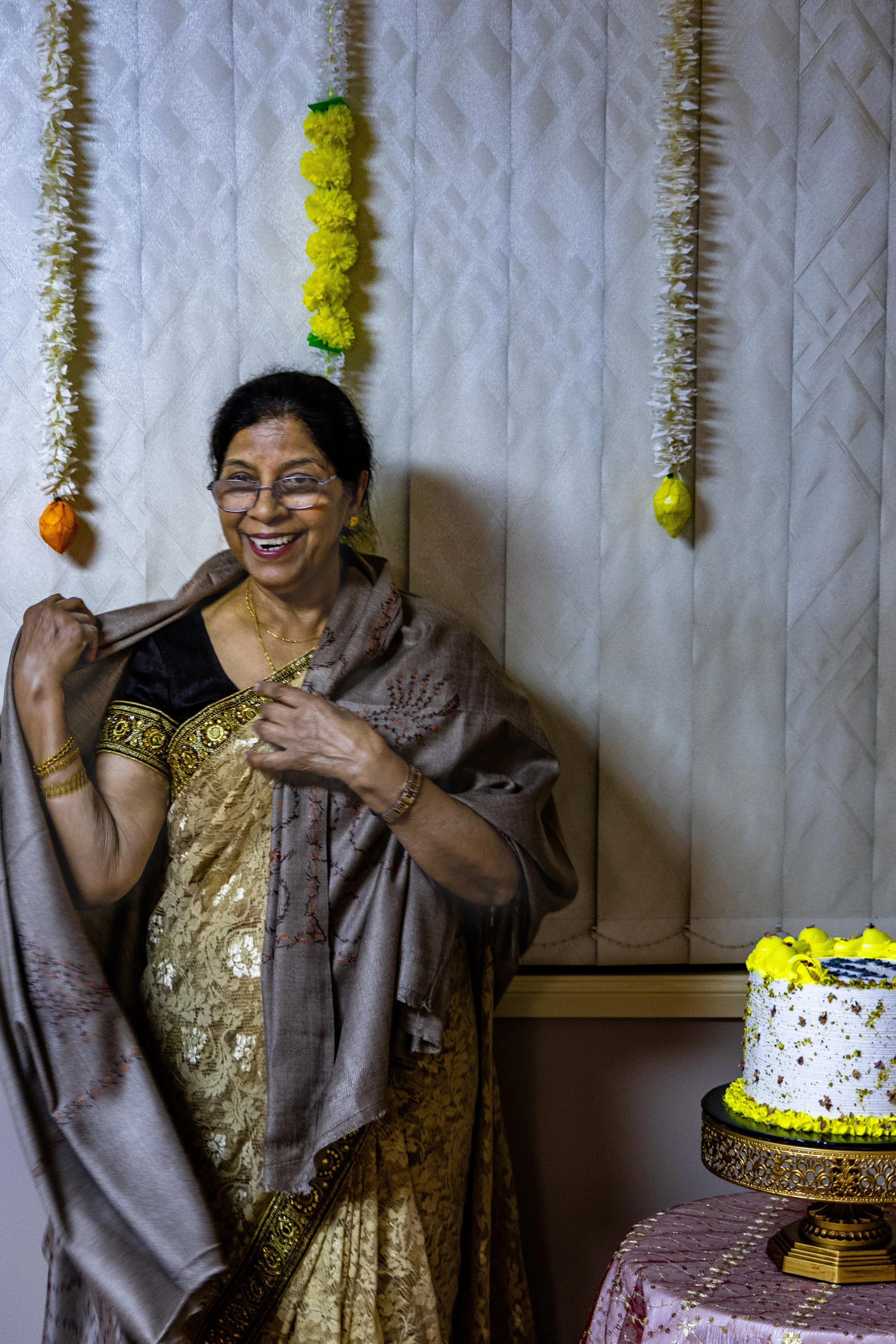 A smiling woman in traditional sari attire standing next to a decorated birthday cake on a table, with colorful hanging decorations in the background.