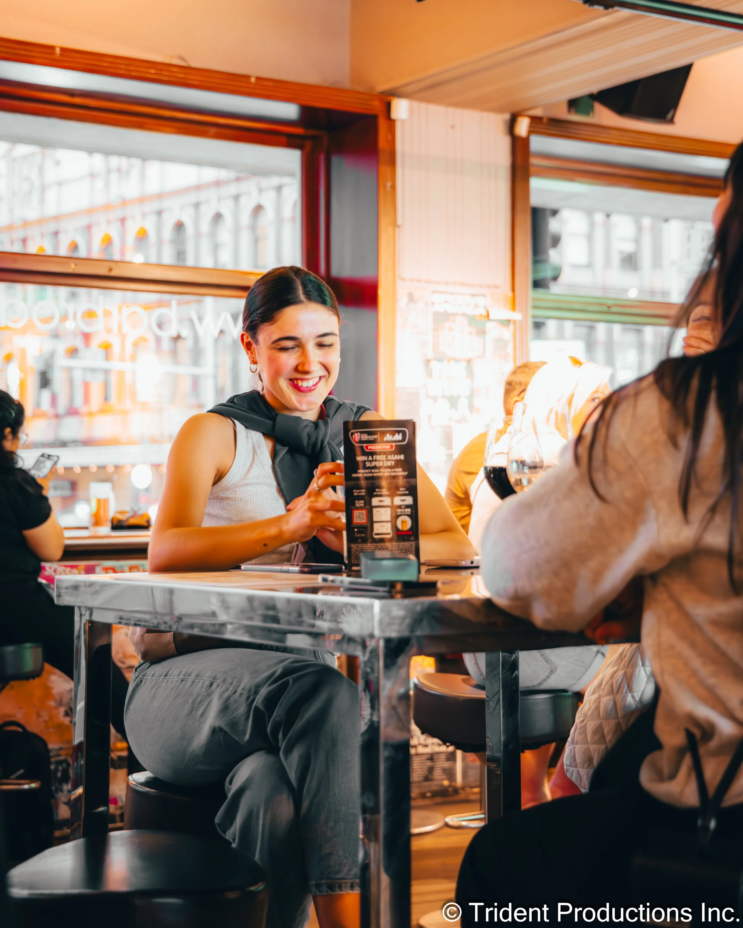 A woman with dark hair tied back, smiling, sitting at a table in a cafe. She is looking at a menu, wearing a sleeveless top and a dark jacket draped over her shoulders. Indoor setting with large windows and a busy city street outside.