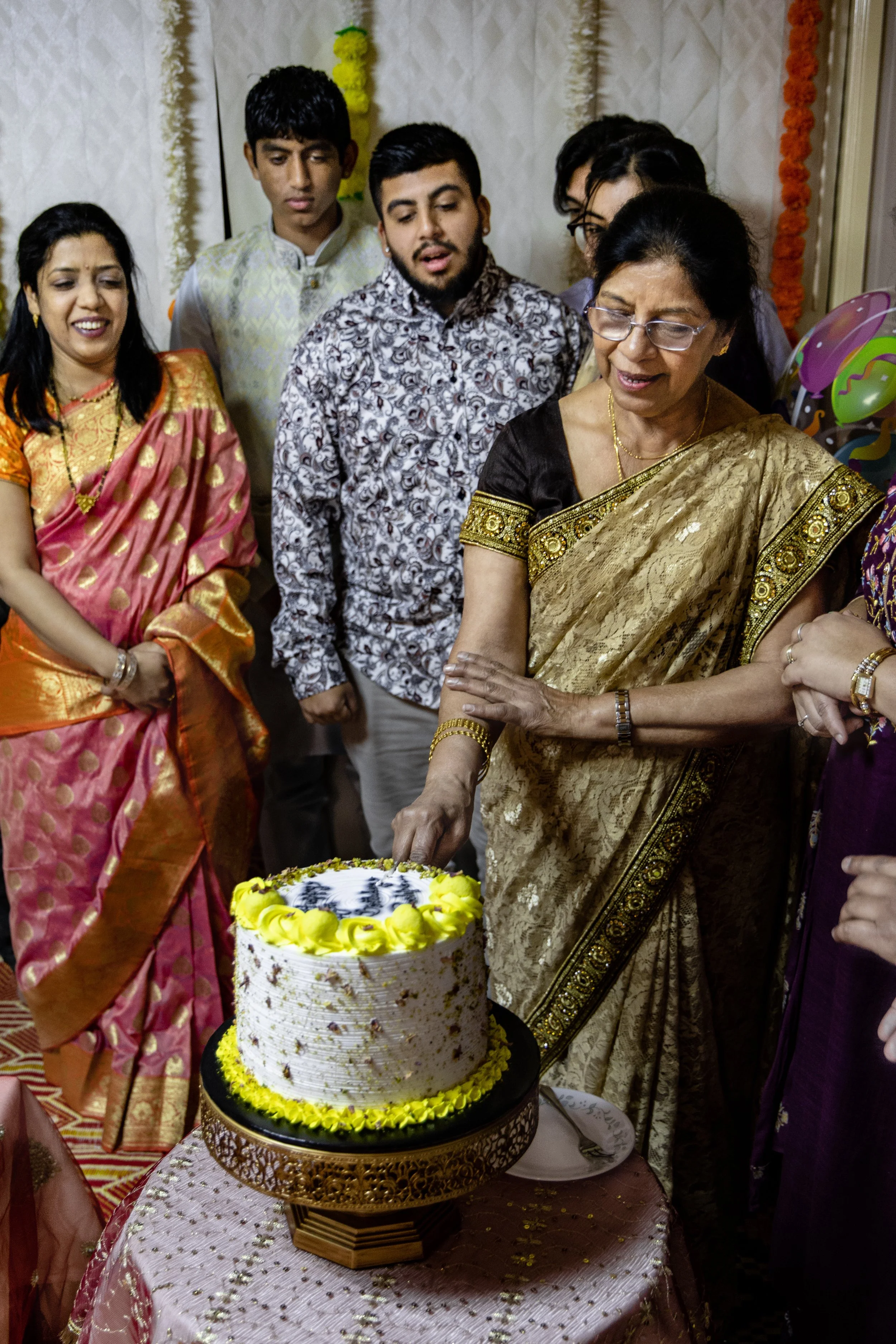 A group of people, including women in traditional Indian sarees and men in dress shirts, gathered around a woman cutting a birthday cake decorated with yellow flowers and a photo on top, at a celebration or party.