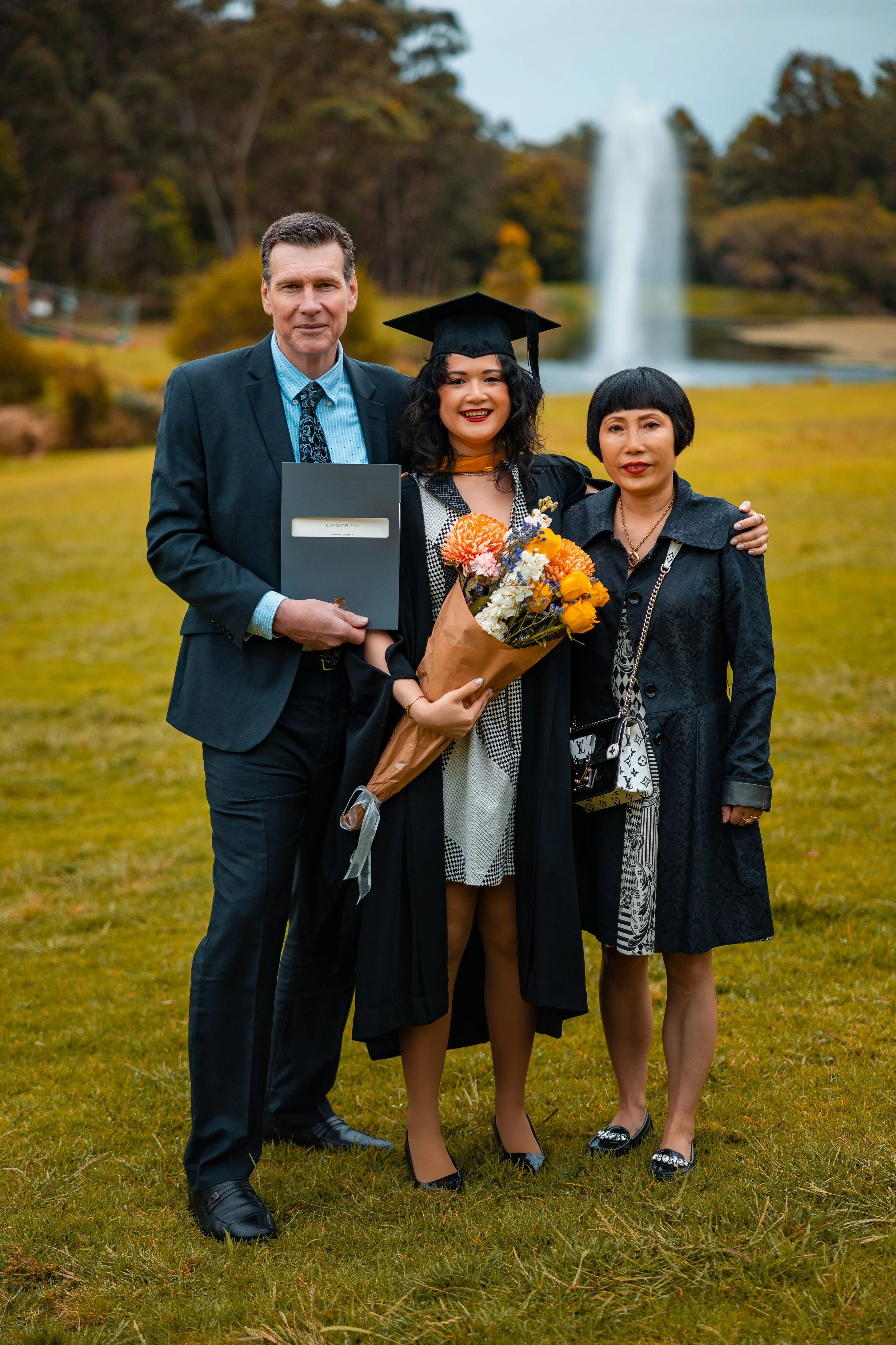 A young woman in a graduation cap and gown holding a bouquet of flowers, standing with her parents outdoors in front of a waterfall.