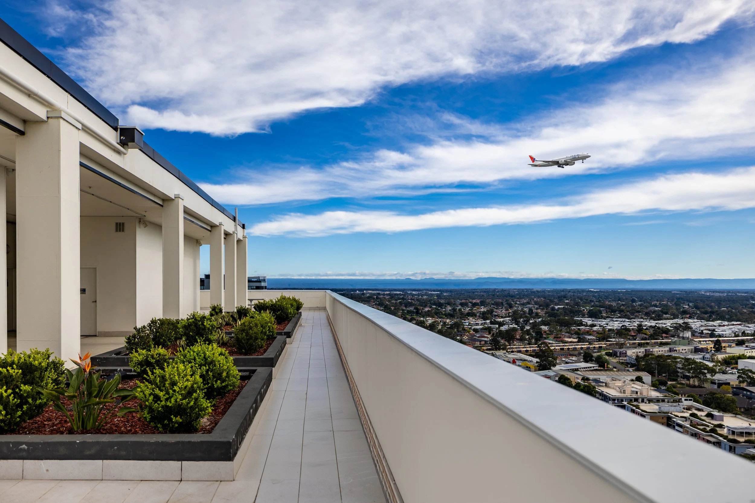View from a rooftop with potted plants and a cityscape in the background, with an airplane flying in a partly cloudy blue sky.