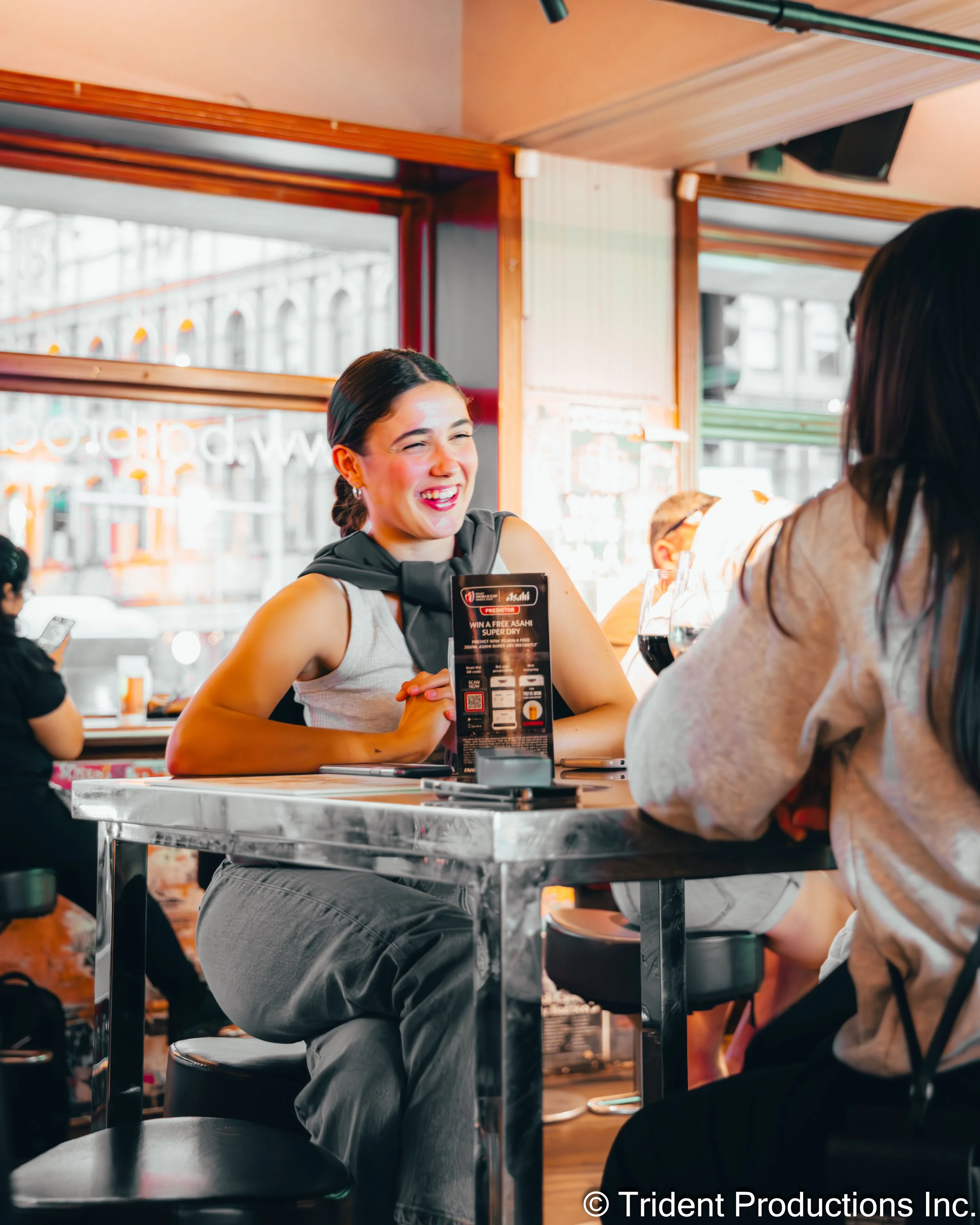 Two women are sitting at a table in a restaurant, smiling and engaged in conversation. One woman has dark hair, tied back, wearing a sleeveless top, and the other has long dark hair, wearing a light-colored hoodie. The table has promotional material 
