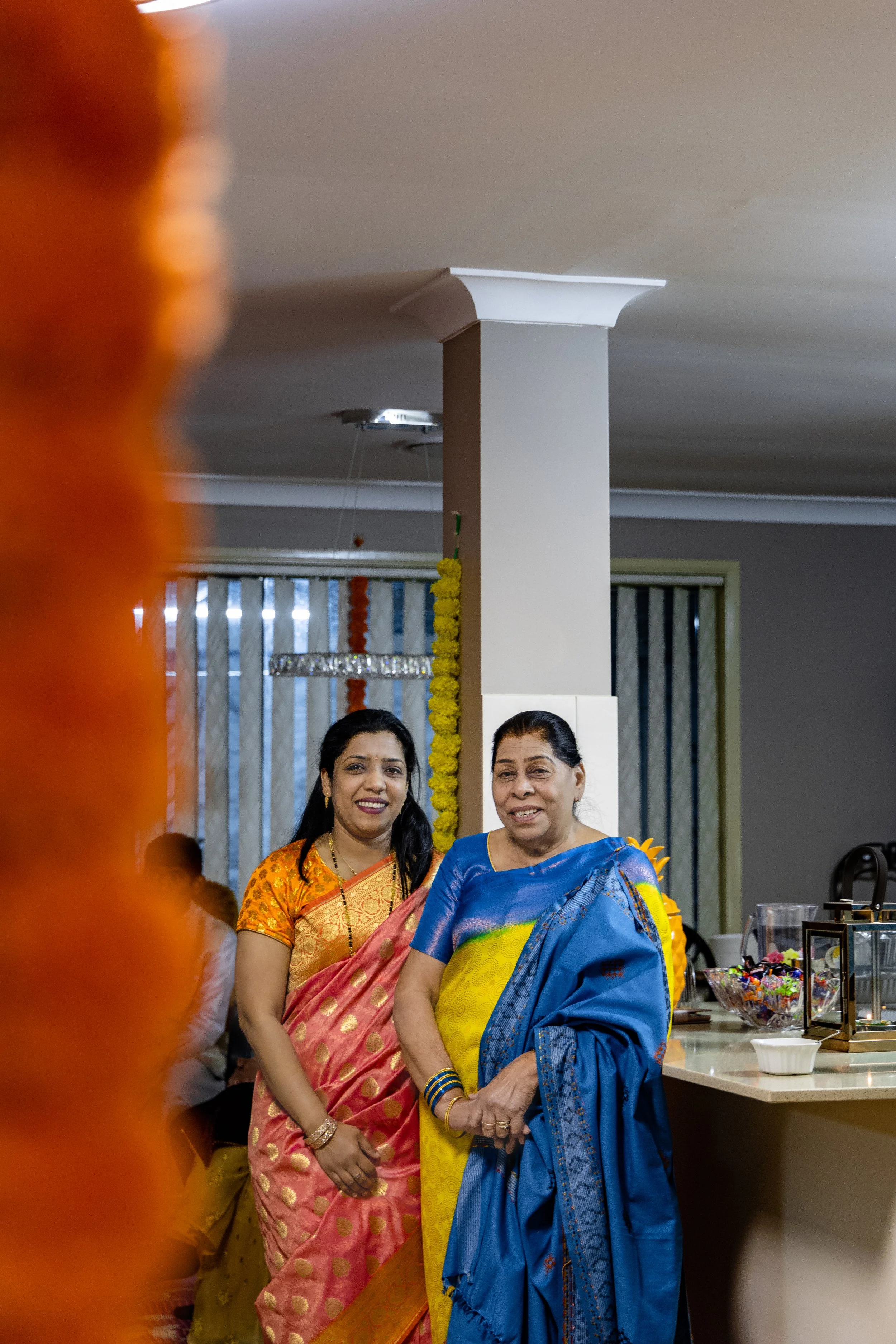 Two women in colorful sarees standing and smiling at a festive gathering, with decorations and food on a counter in the background.