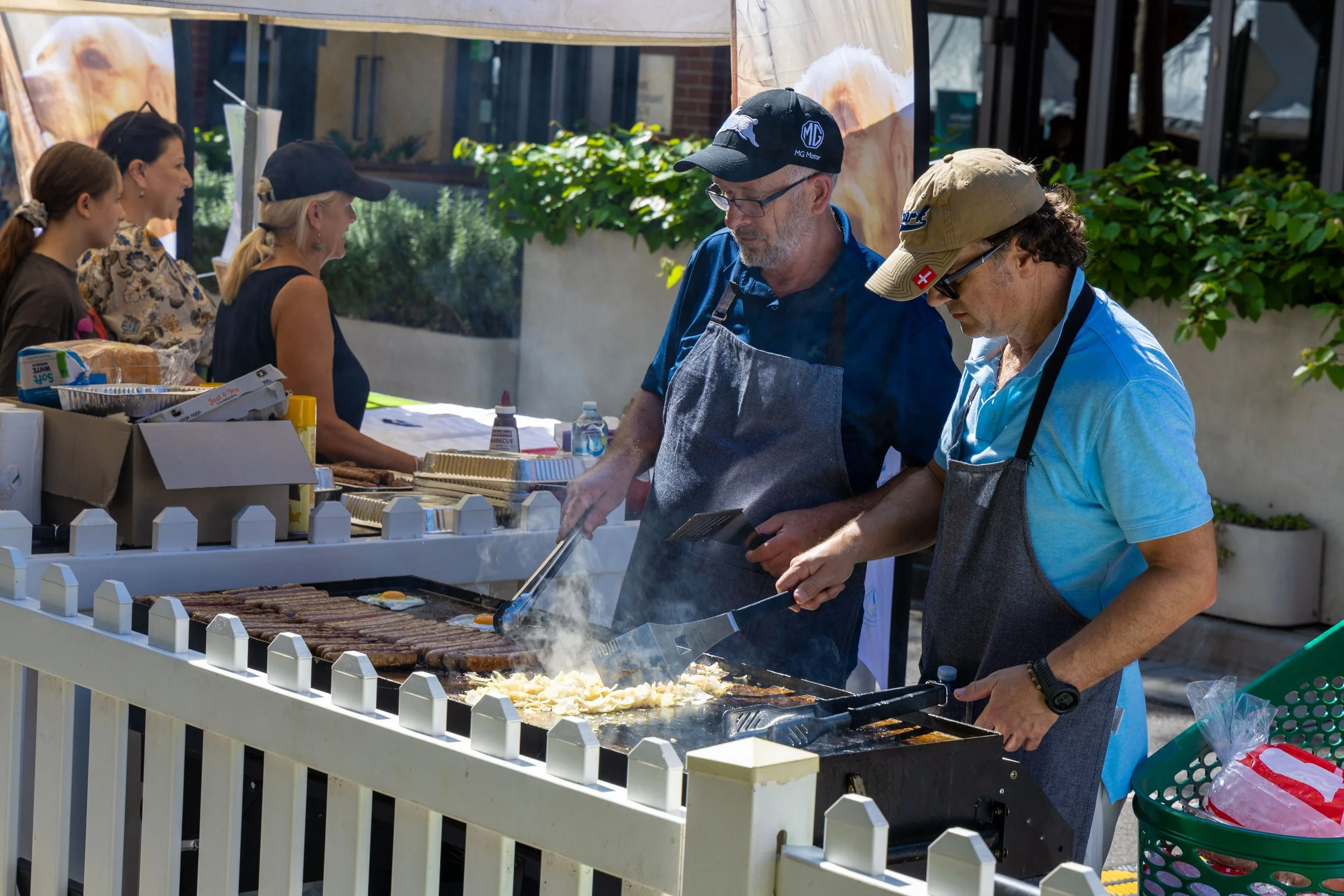 Two men wearing aprons and hats cook steak and another food item on a large outdoor grill during a barbecue event, with women and food supplies seen in the background.