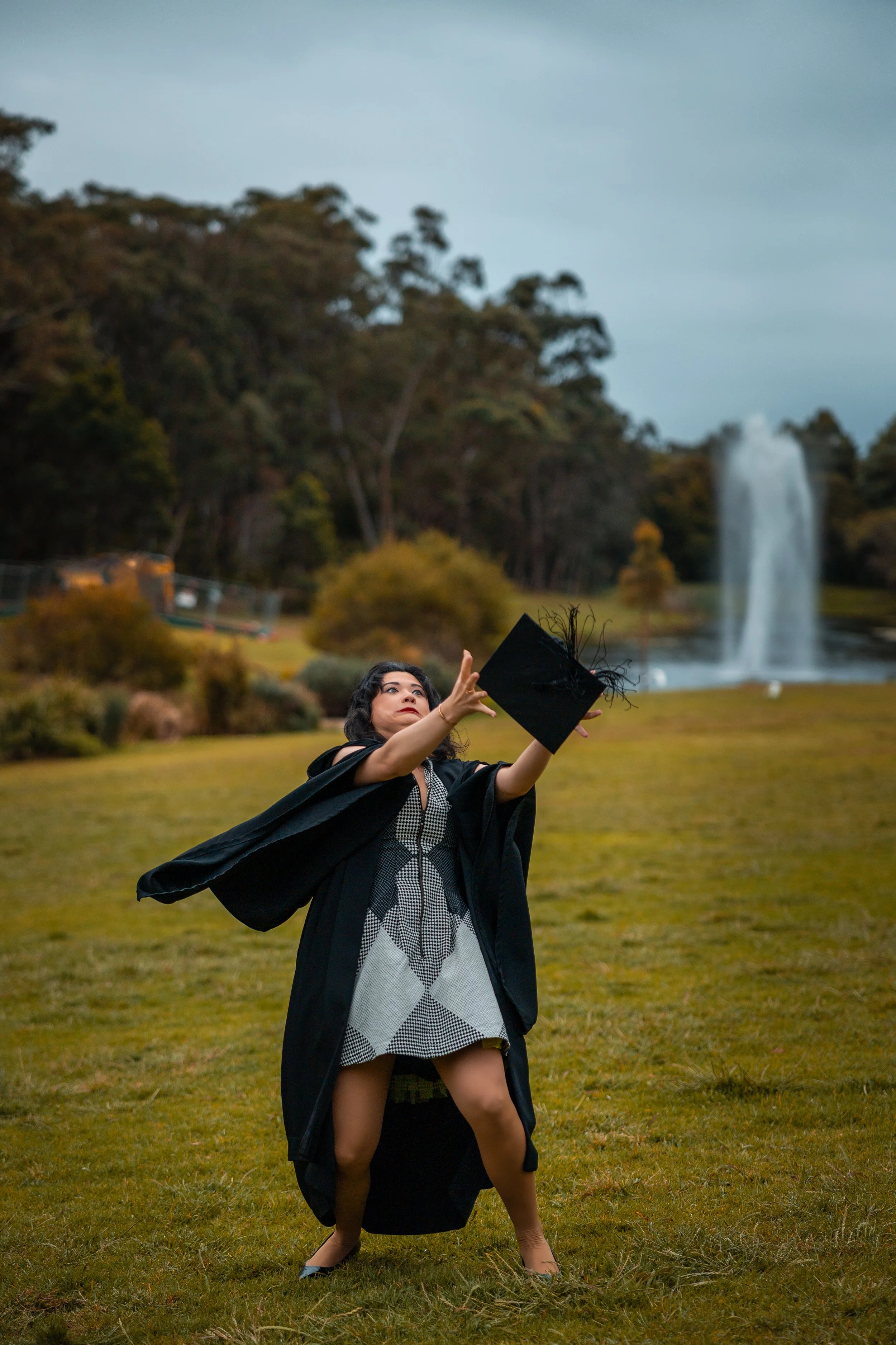 A woman in a graduation gown holding a cap in a grassy area with a waterfall in the background.