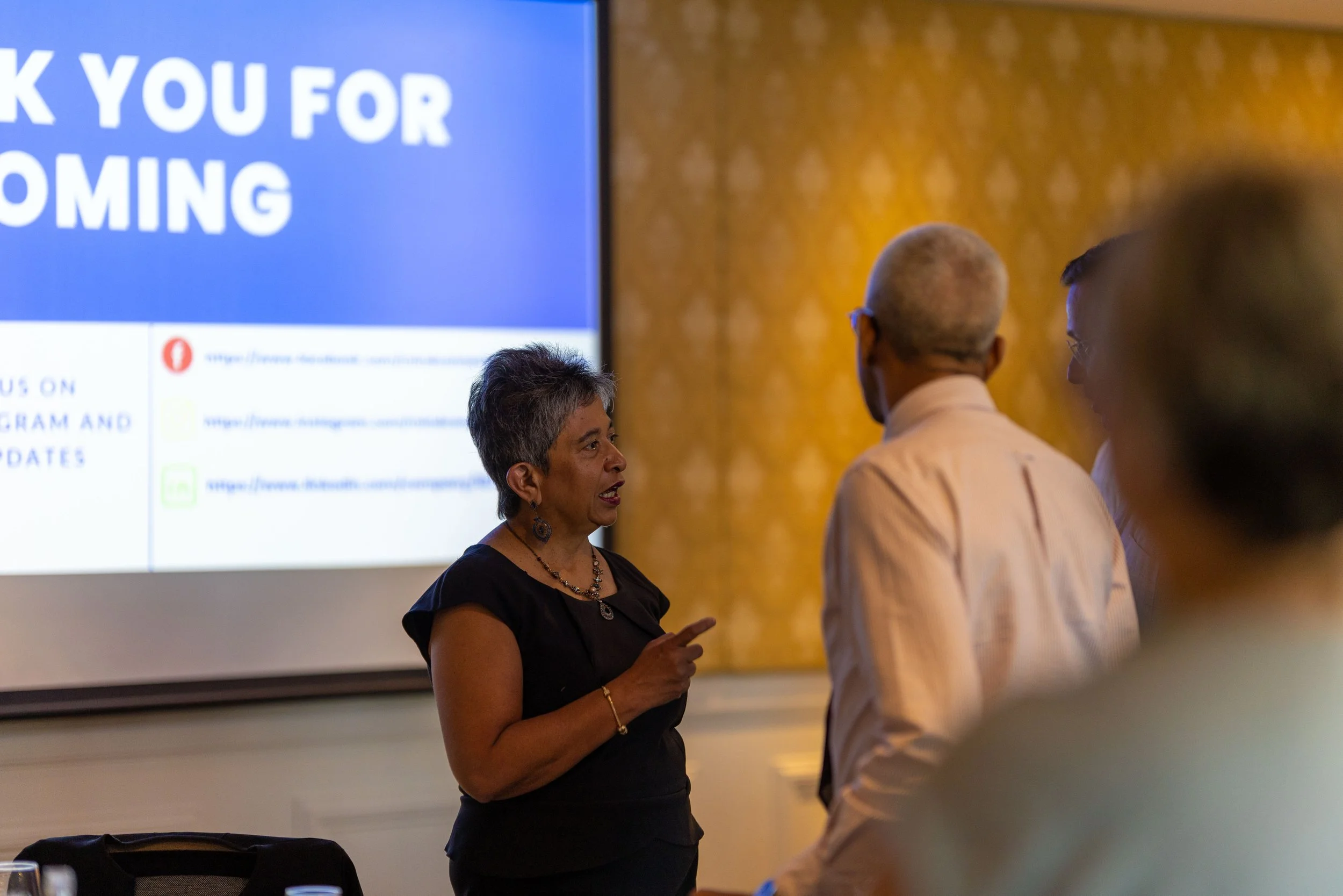 A woman with short gray hair talking to a group of people in a room with a large screen displaying a 'Thank You for Coming' message.