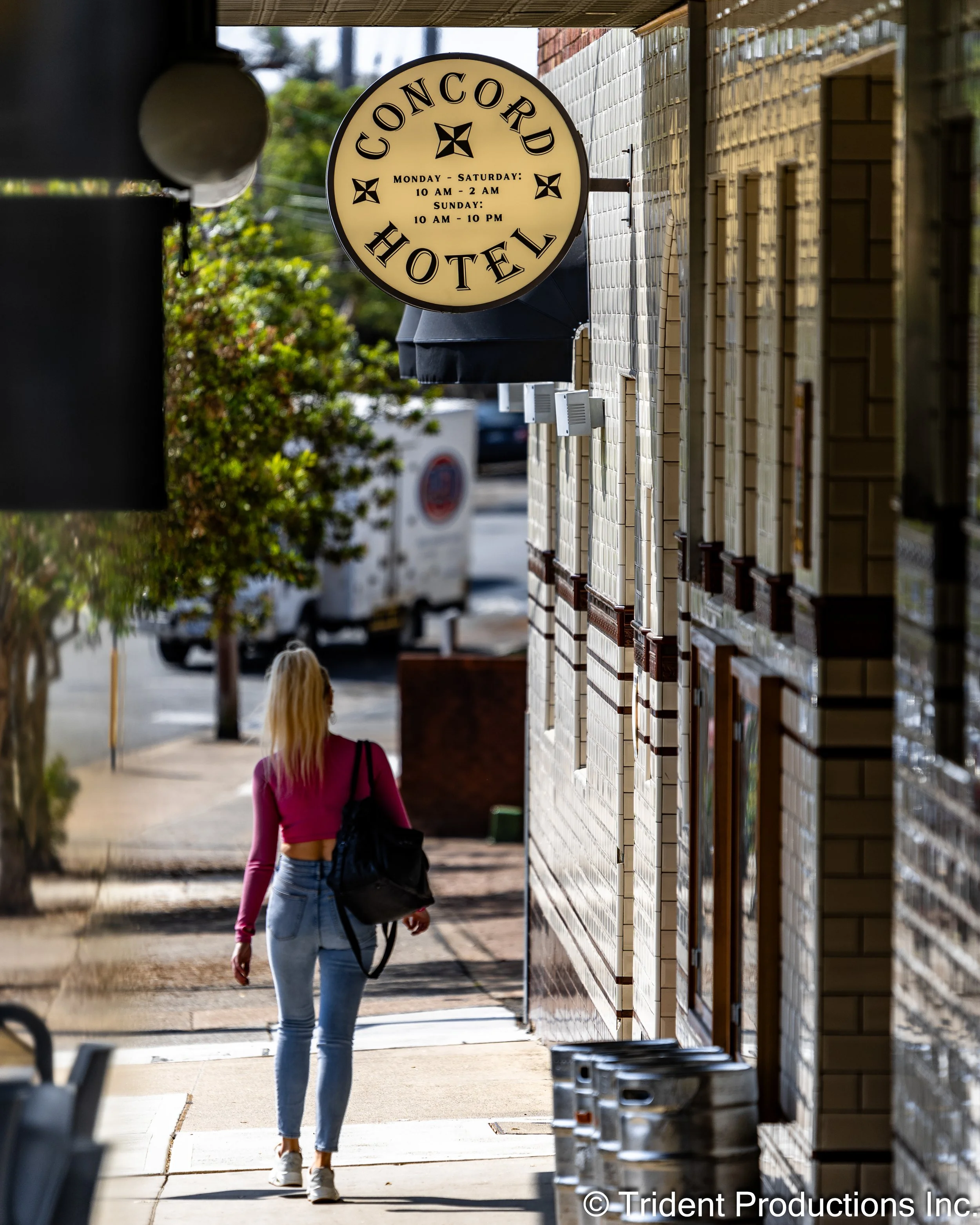 A woman walking on sidewalk outside a building with a sign for Concord Hotel, showing hours of operation.