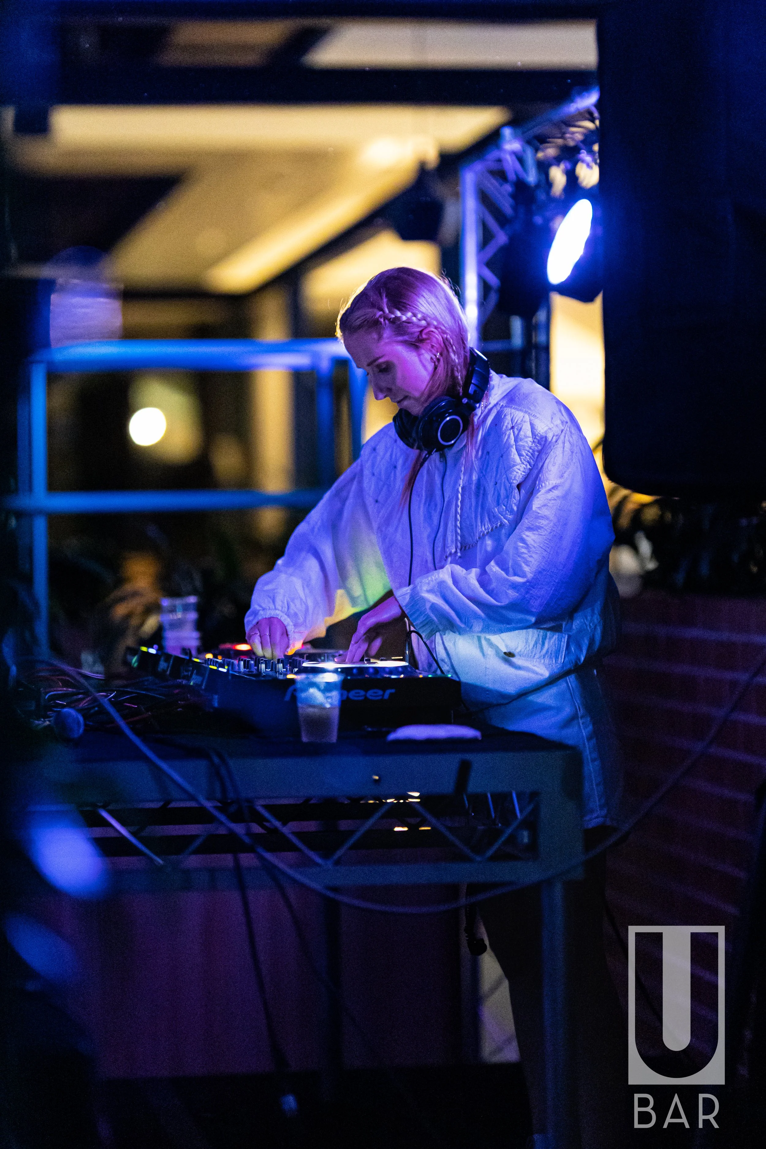 A female DJ with short blonde hair, wearing a light-colored jacket and headphones, performing at a dimly lit nightclub with purple and blue lighting. She is adjusting controls on a DJ mixer.