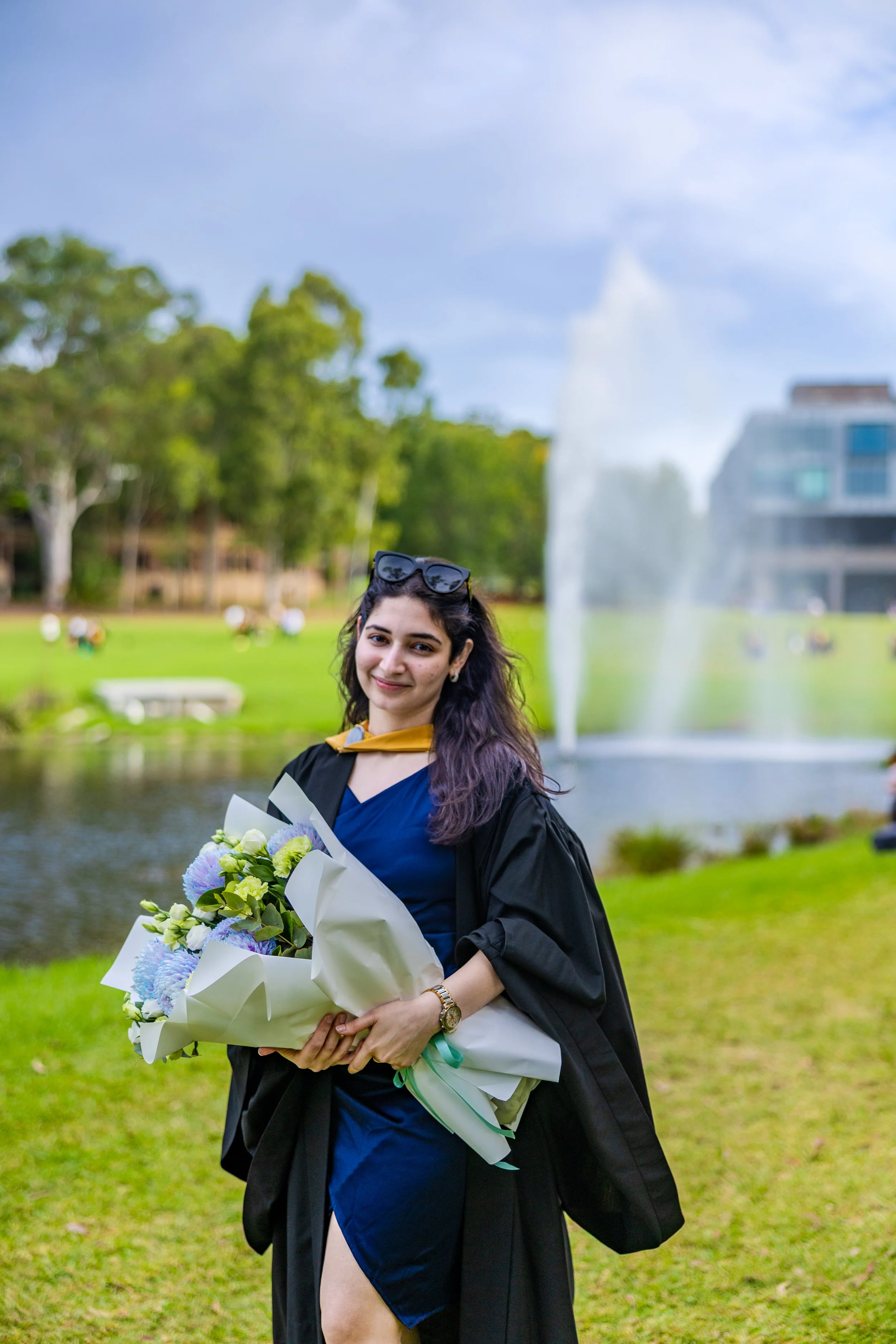 A young woman in a graduation gown holding a bouquet of flowers outdoors near a fountain on a sunny day.