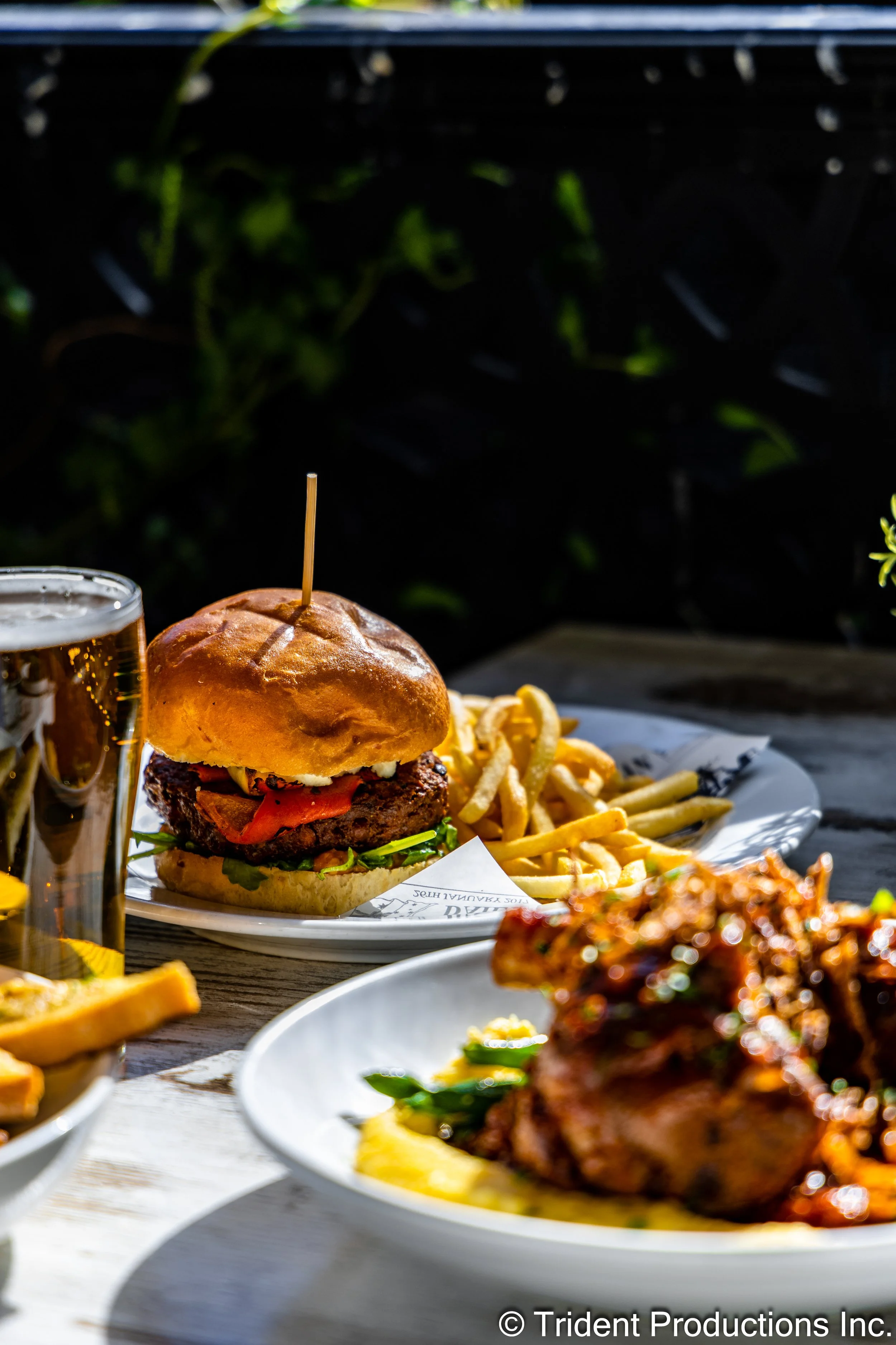 A plate with a cheeseburger, French fries, and a glass of beer on a wooden table outdoors.