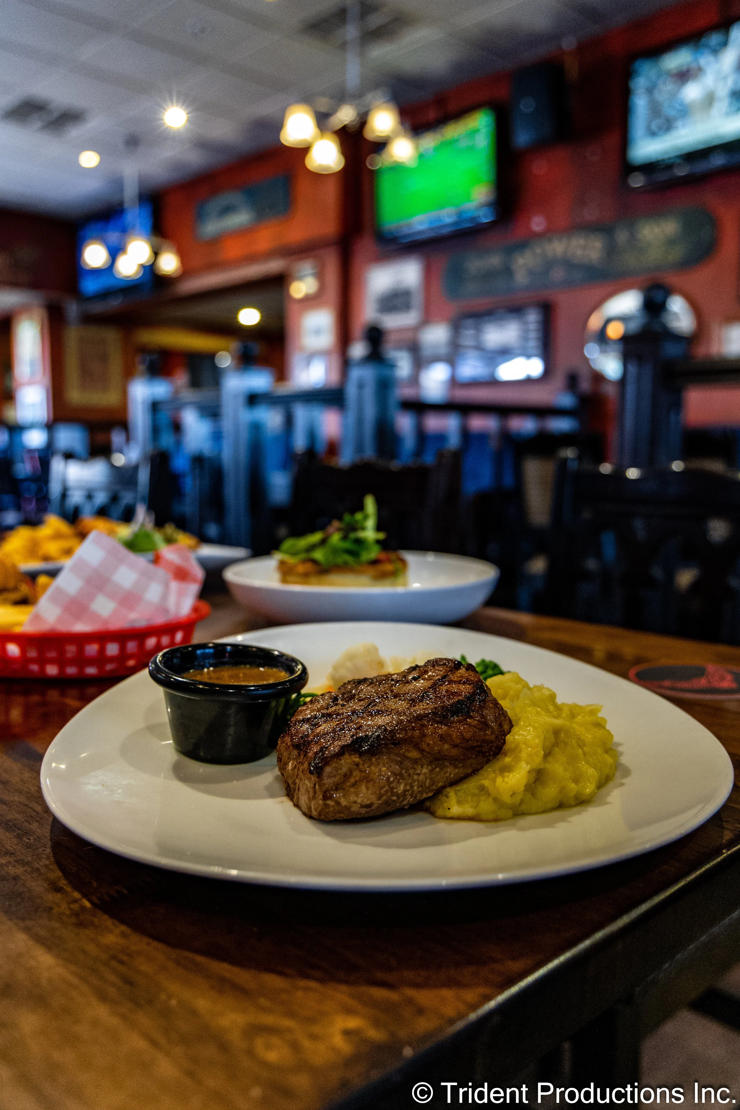 A plate with grilled steak, mashed potatoes, and a small container of sauce on a wooden table inside a restaurant.