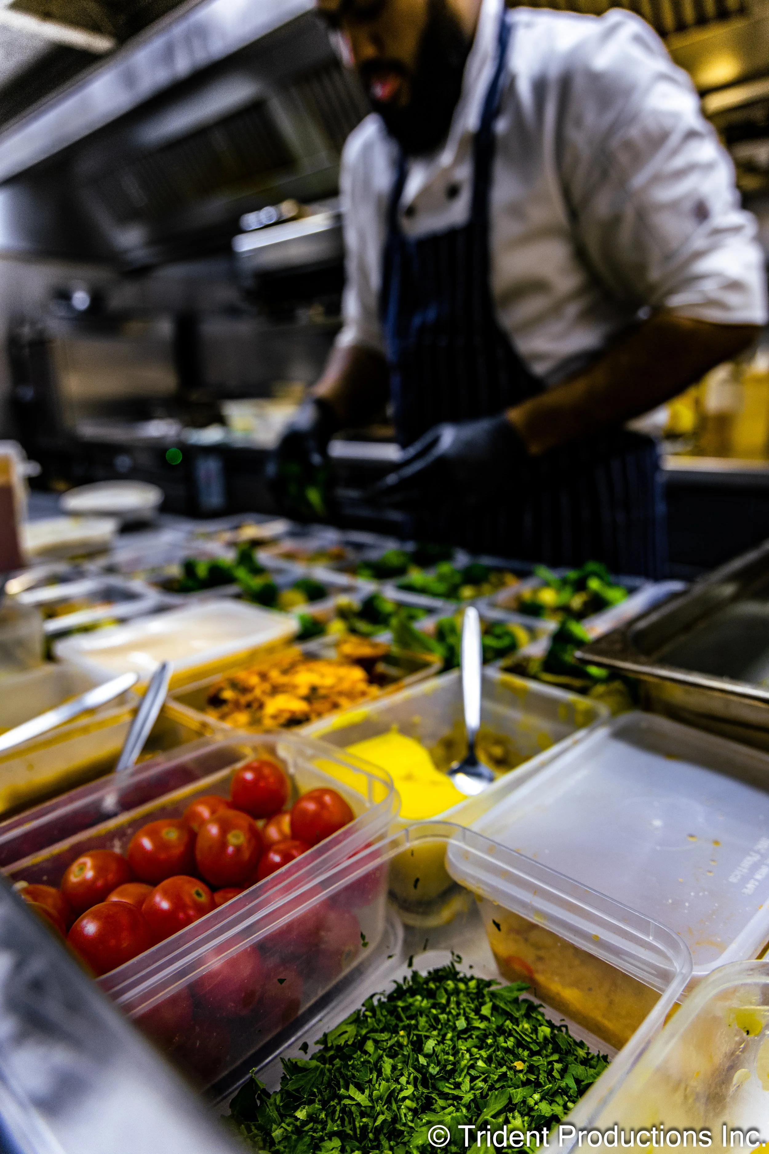 Chef preparing vegetables behind a counter with cherry tomatoes, chopped herbs, and various sauces or dressings in containers.