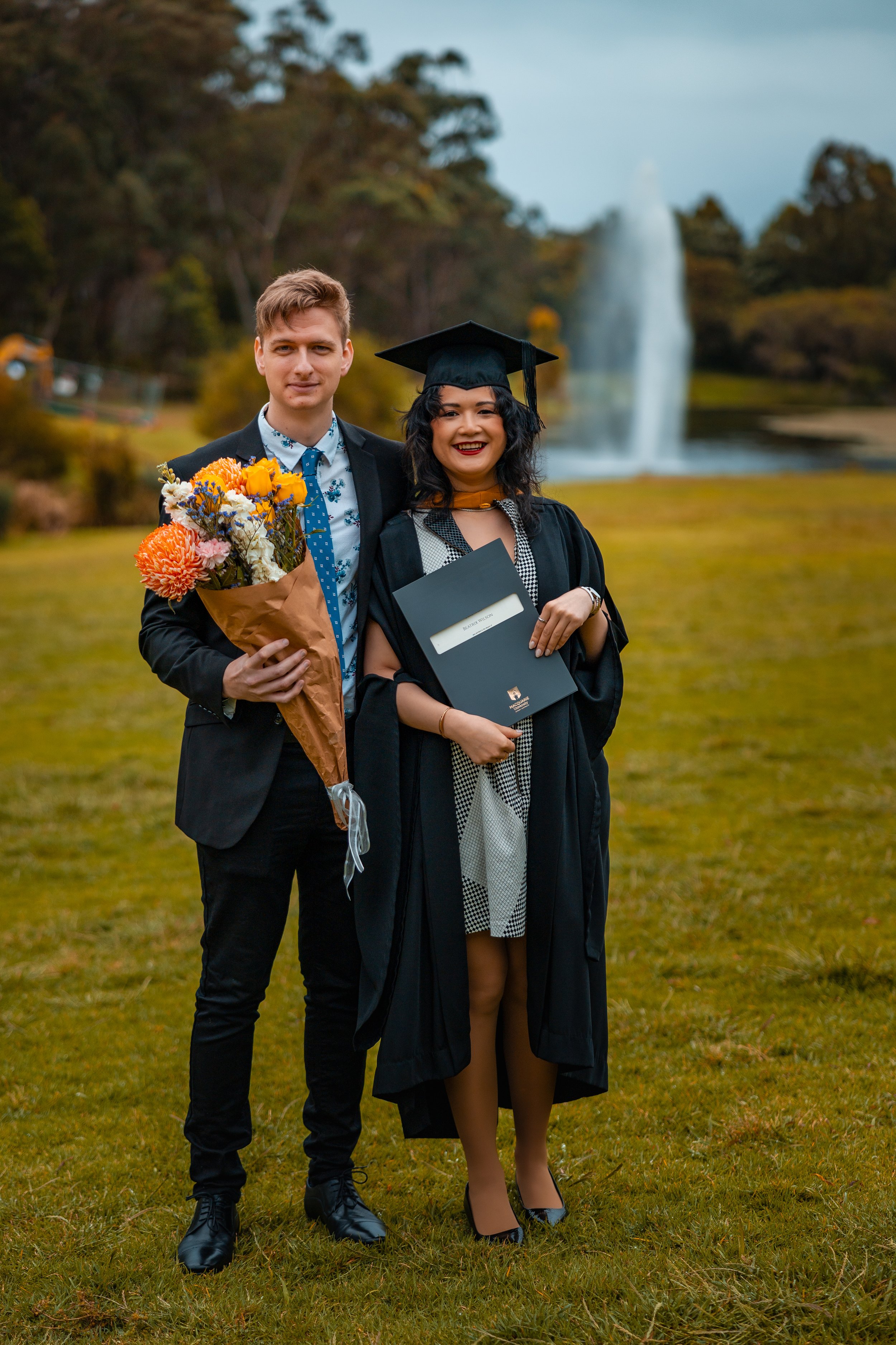 Two people in graduation attire, standing on a grassy field, holding a bouquet of flowers and a diploma, with a waterfall and trees in the background.