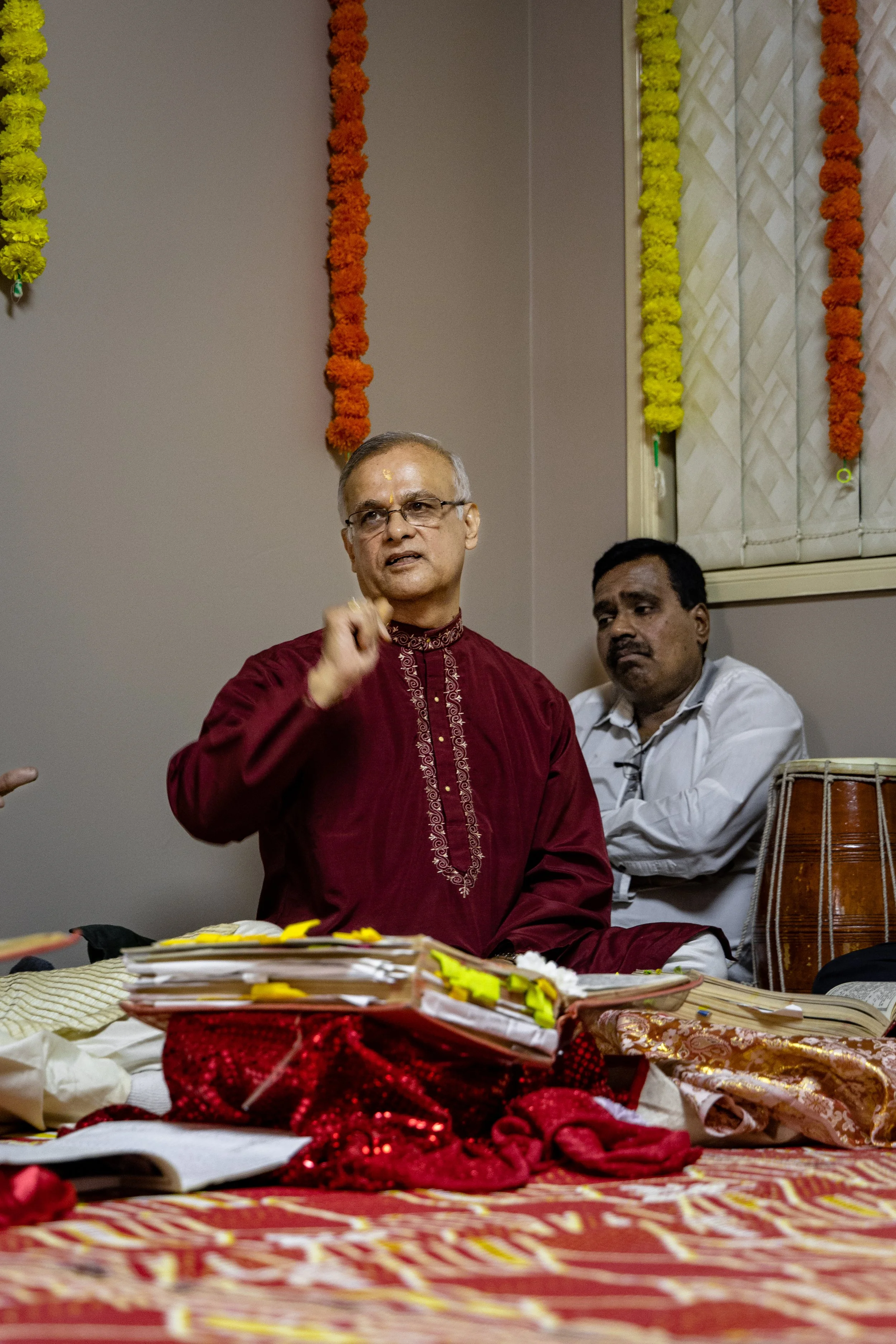 A man in a traditional red Indian kurta speaking or explaining during a ceremony or celebration, sitting on a decorated surface with papers, books, and cloth laid out in front of him. Another man in white is sitting behind him, listening. The backgro