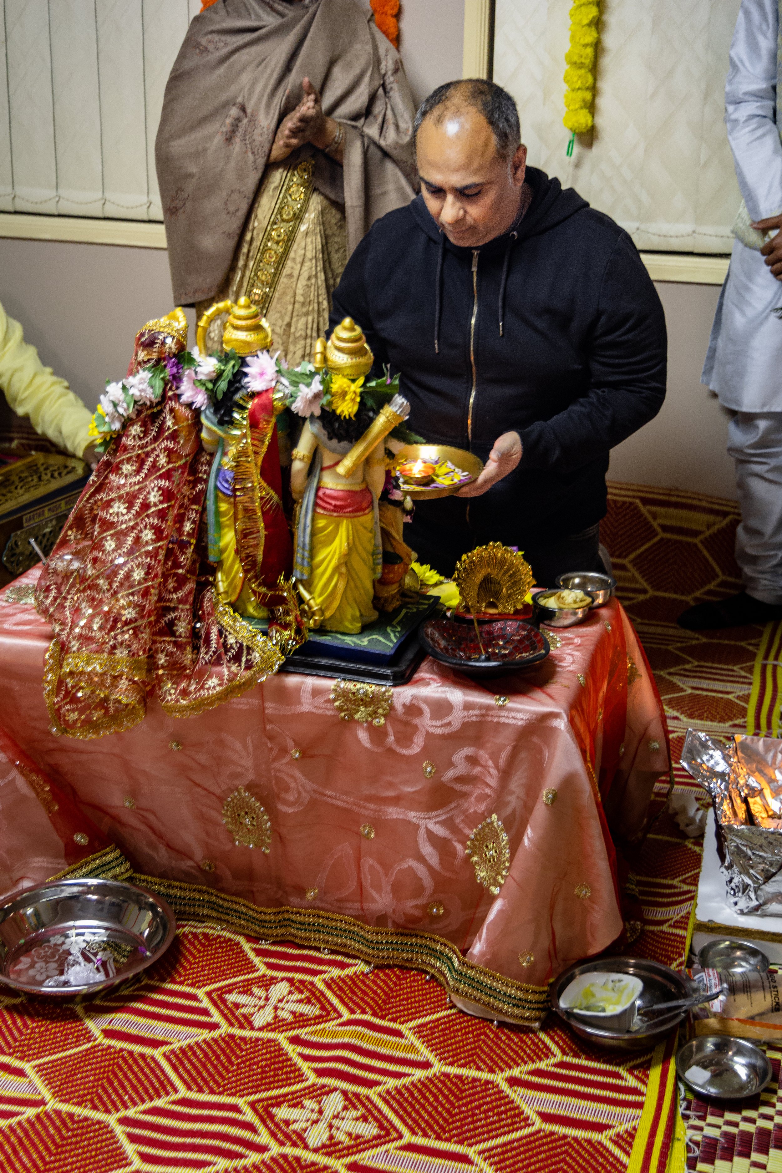 Man in black hoodie performing a religious ritual in front of decorated Hindu idols and statues on a pink table covered with a pink cloth, with offerings and incense, surrounded by other worshippers.