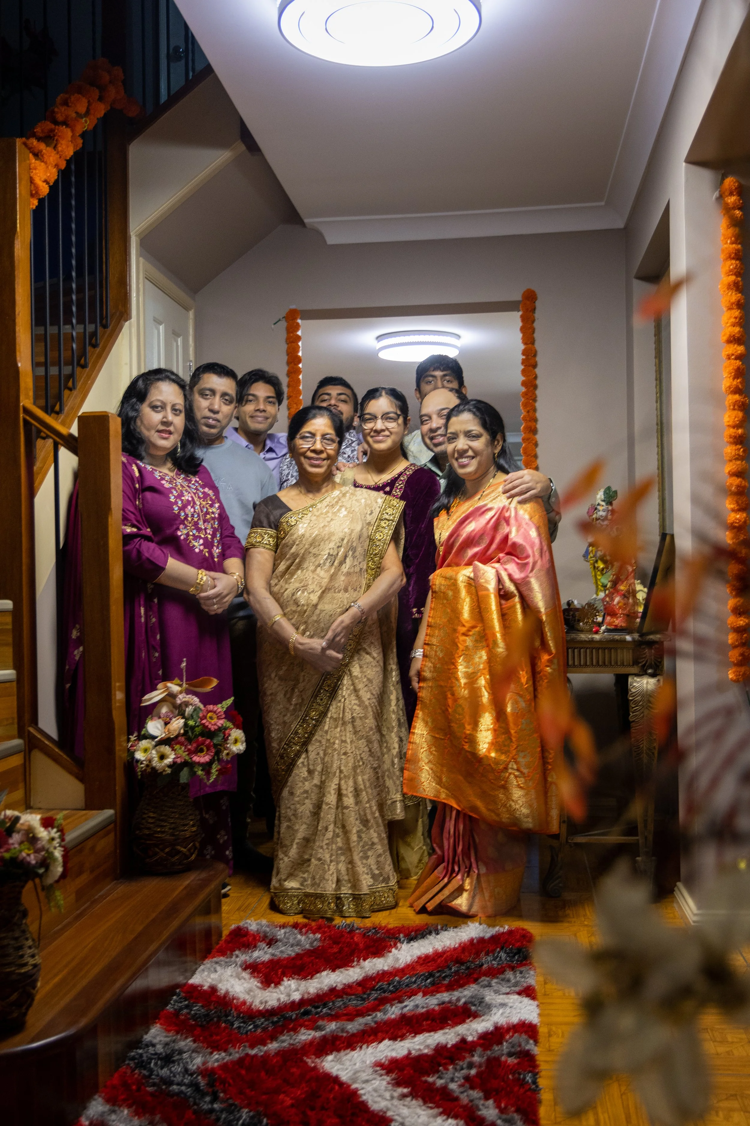 Group of people celebrating a family event in a decorated home, dressed in traditional Indian sarees and clothes, standing together smiling on a stairway with flower garlands and floral decorations.
