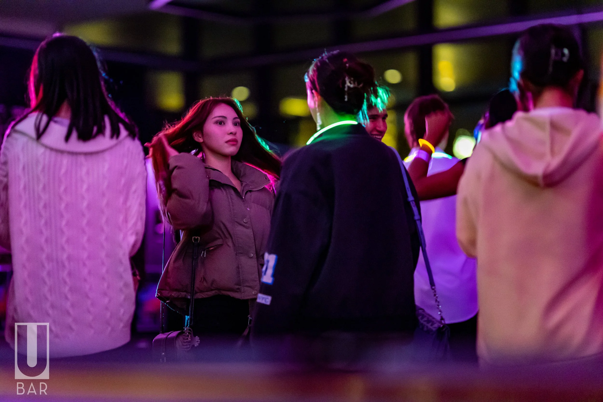 Group of people in a dimly lit bar or club with colorful neon lighting, engaged in conversation and enjoying the nightlife scene, featuring women and men, some with glow bracelets.