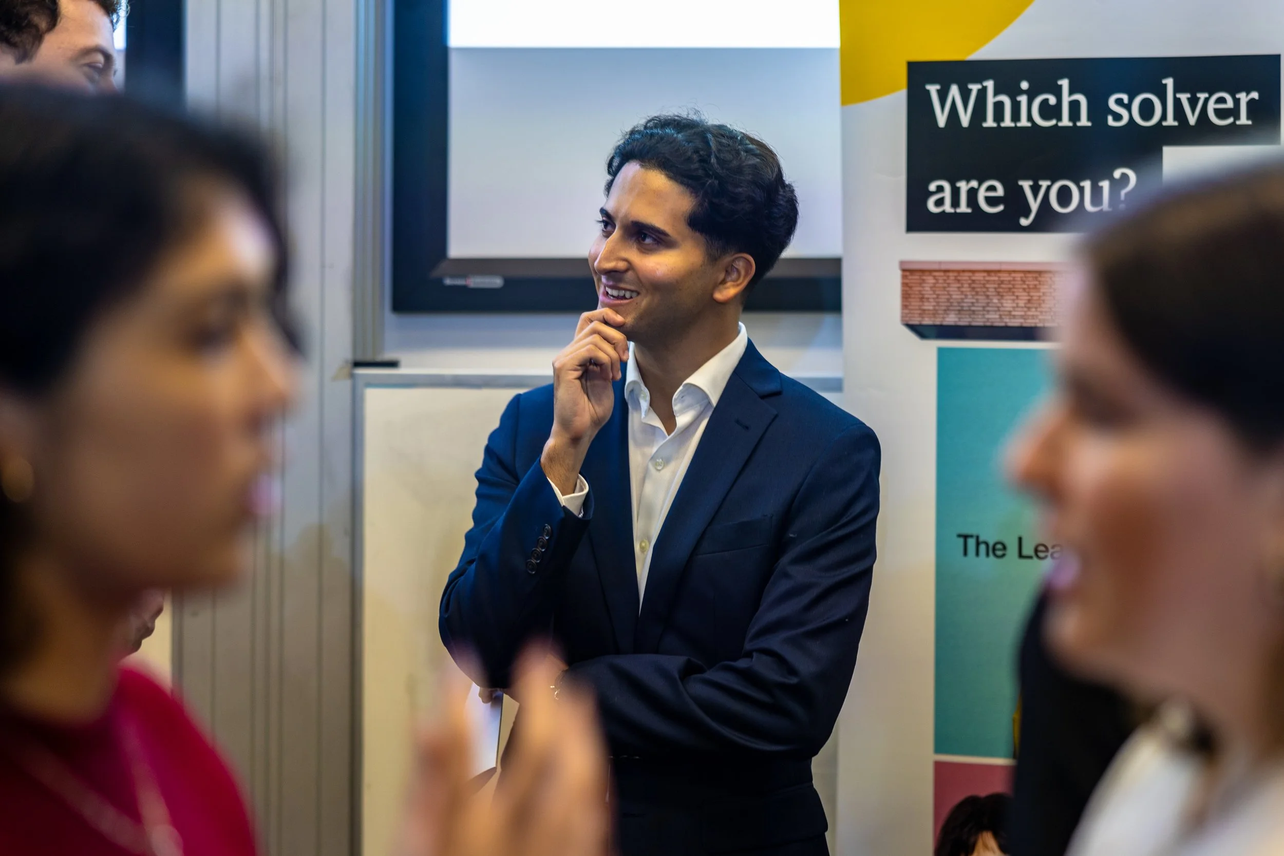 A man in a navy suit and white shirt is standing with his hand on his chin, smiling thoughtfully at a group of people in a professional setting. The background shows a banner with the question 'Which solver are you?' and parts of a presentation or di