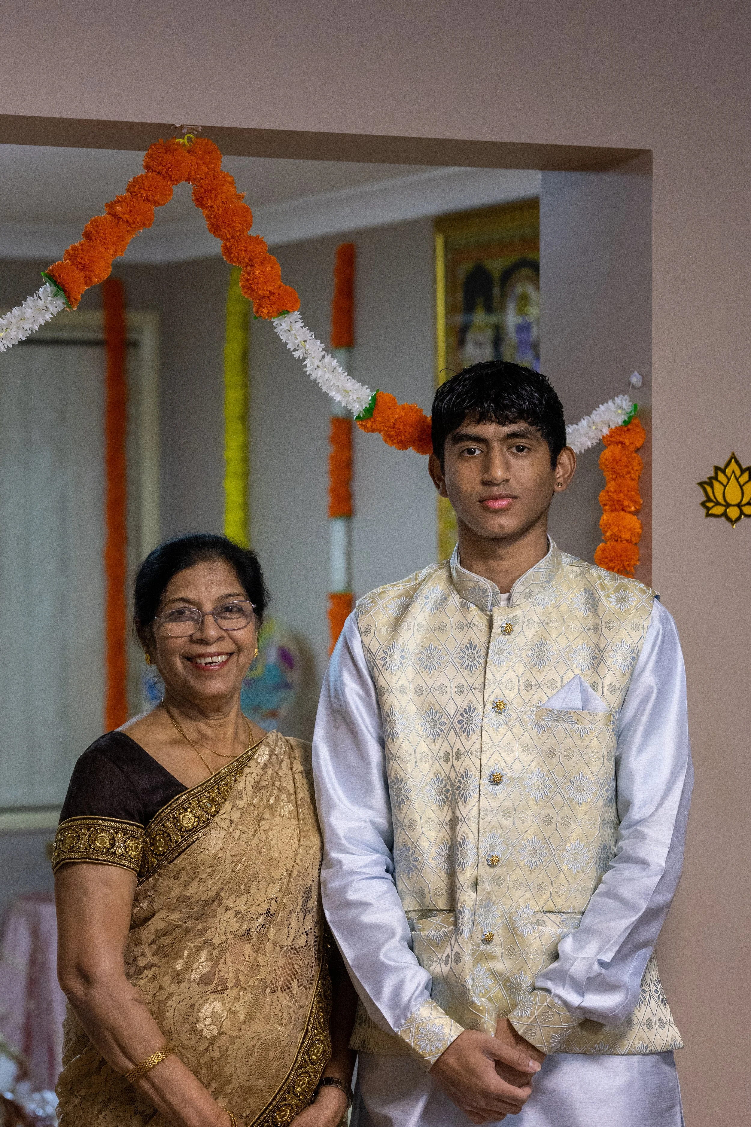 A young man and an older woman dressed in traditional Indian attire standing together at a decorated indoor event, with marigold flower garlands and other decorations visible in the background.