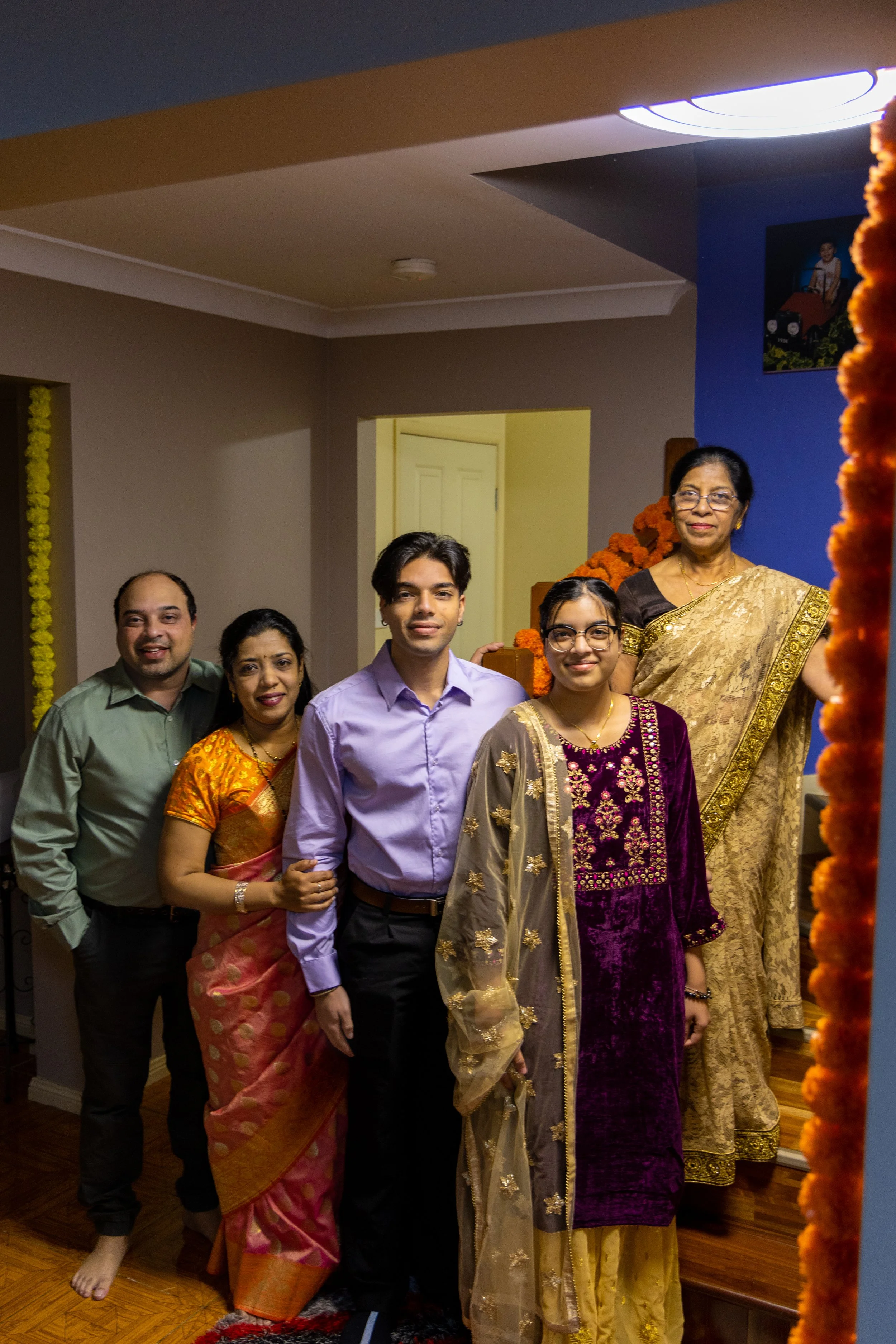 Group of five people standing in a living room with a woman on the stairs, dressed in traditional Indian attire, celebrating a cultural event.