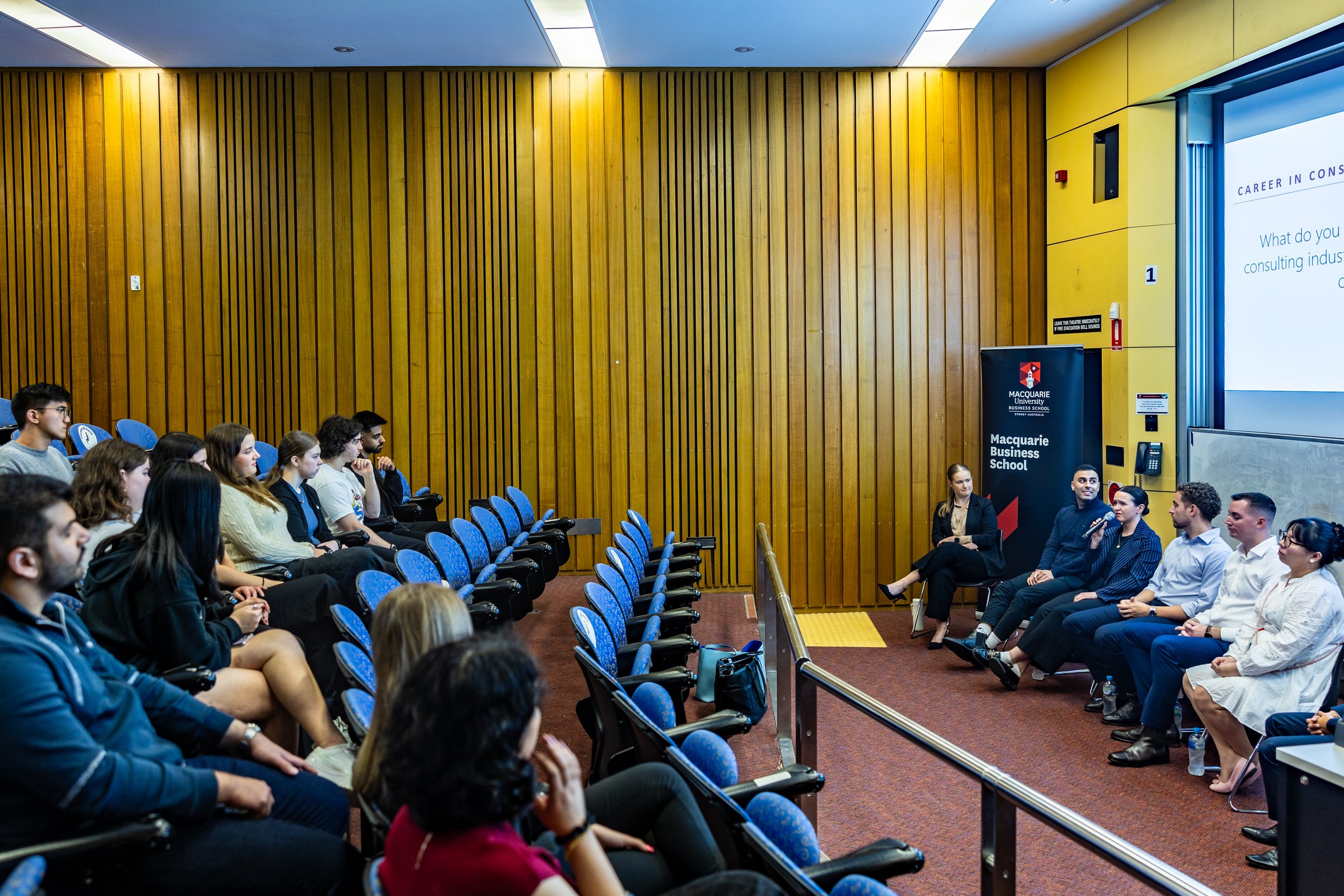 Students attending a panel discussion in a university lecture hall with wooden panel walls, blue seats, and a Macquarie Business School banner.