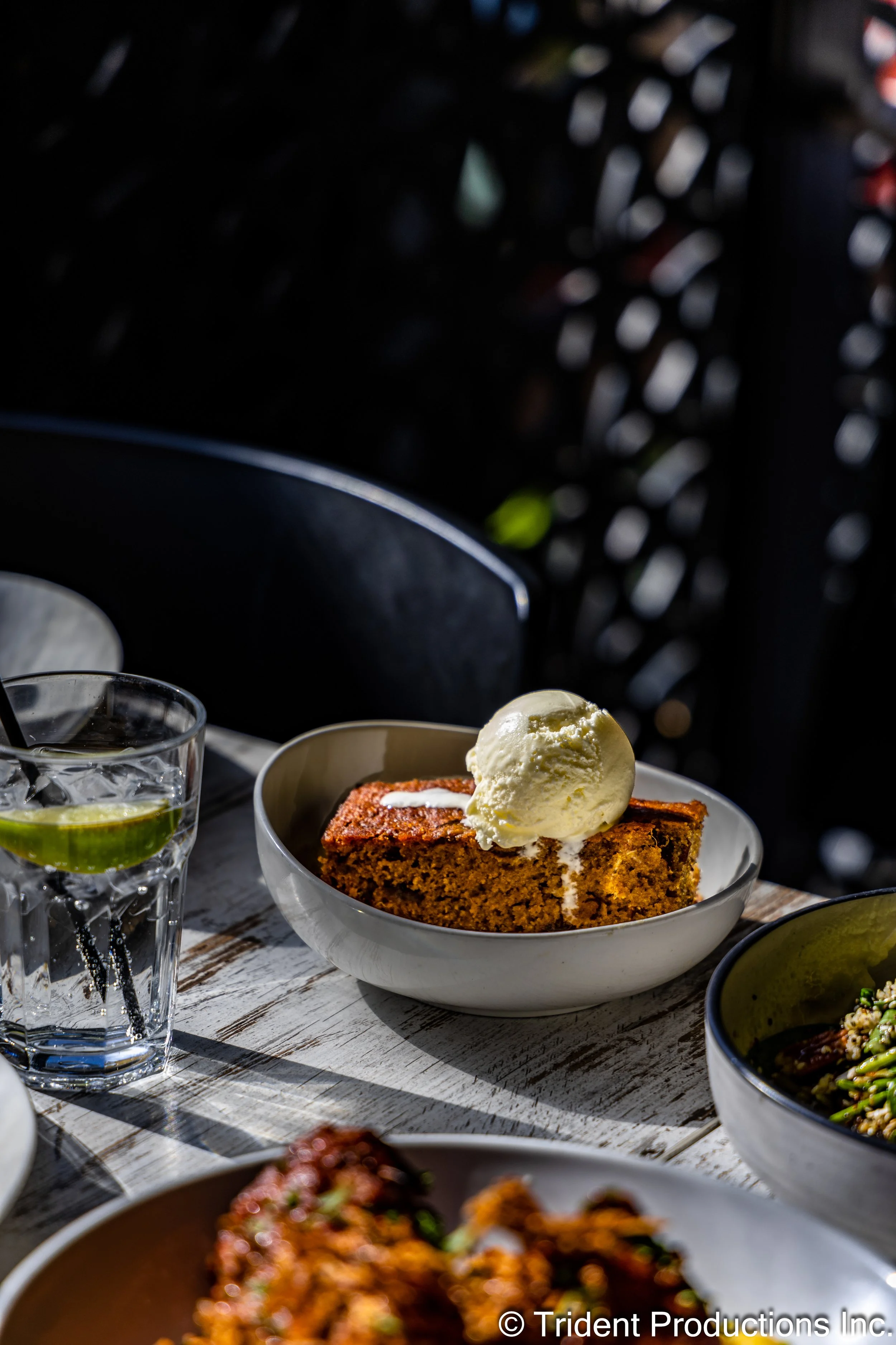 A slice of carrot cake with a scoop of vanilla ice cream on top, served in a white bowl, on a rustic wooden table.