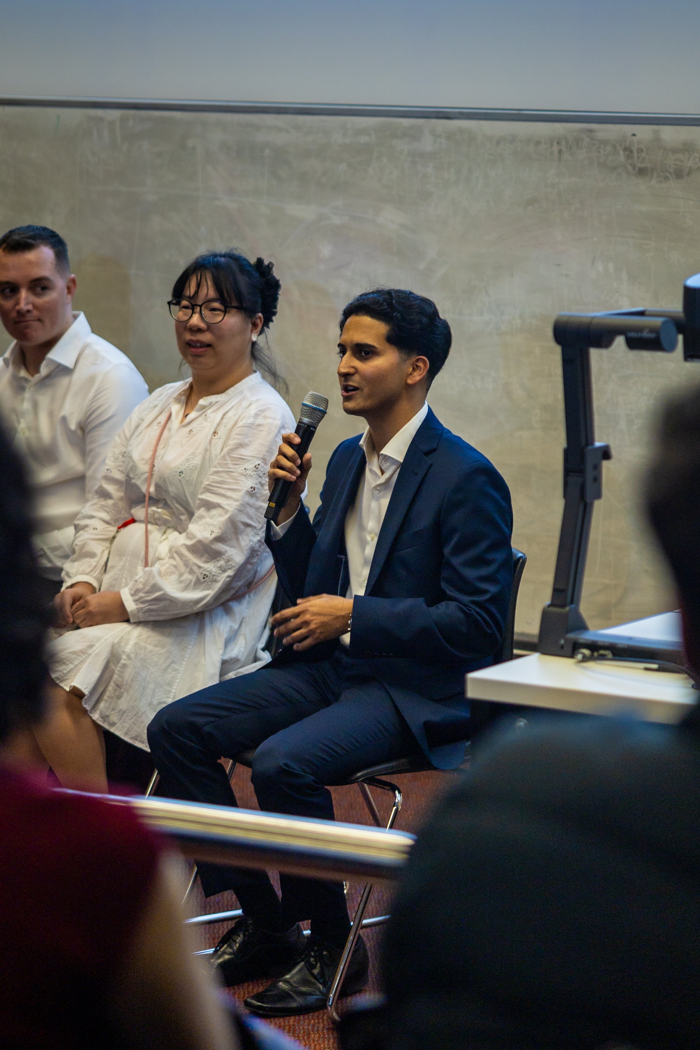 A man in a navy suit holding a microphone participating in a panel discussion, seated next to women in white attire, in a classroom or conference room setting.