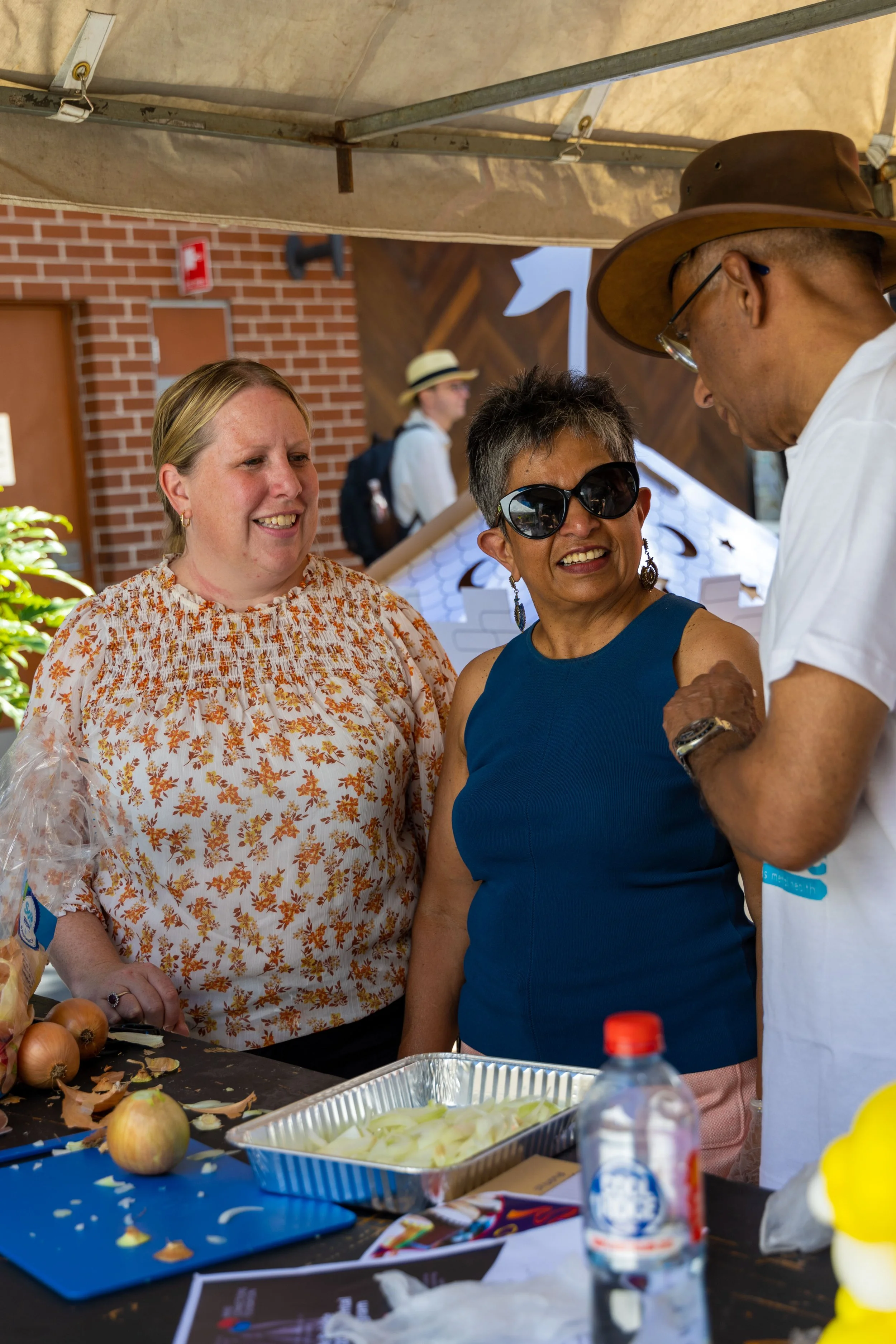 Three women and one man talking at an outdoor event, with a table of onions, onions peelings, and a foil tray in front of them.
