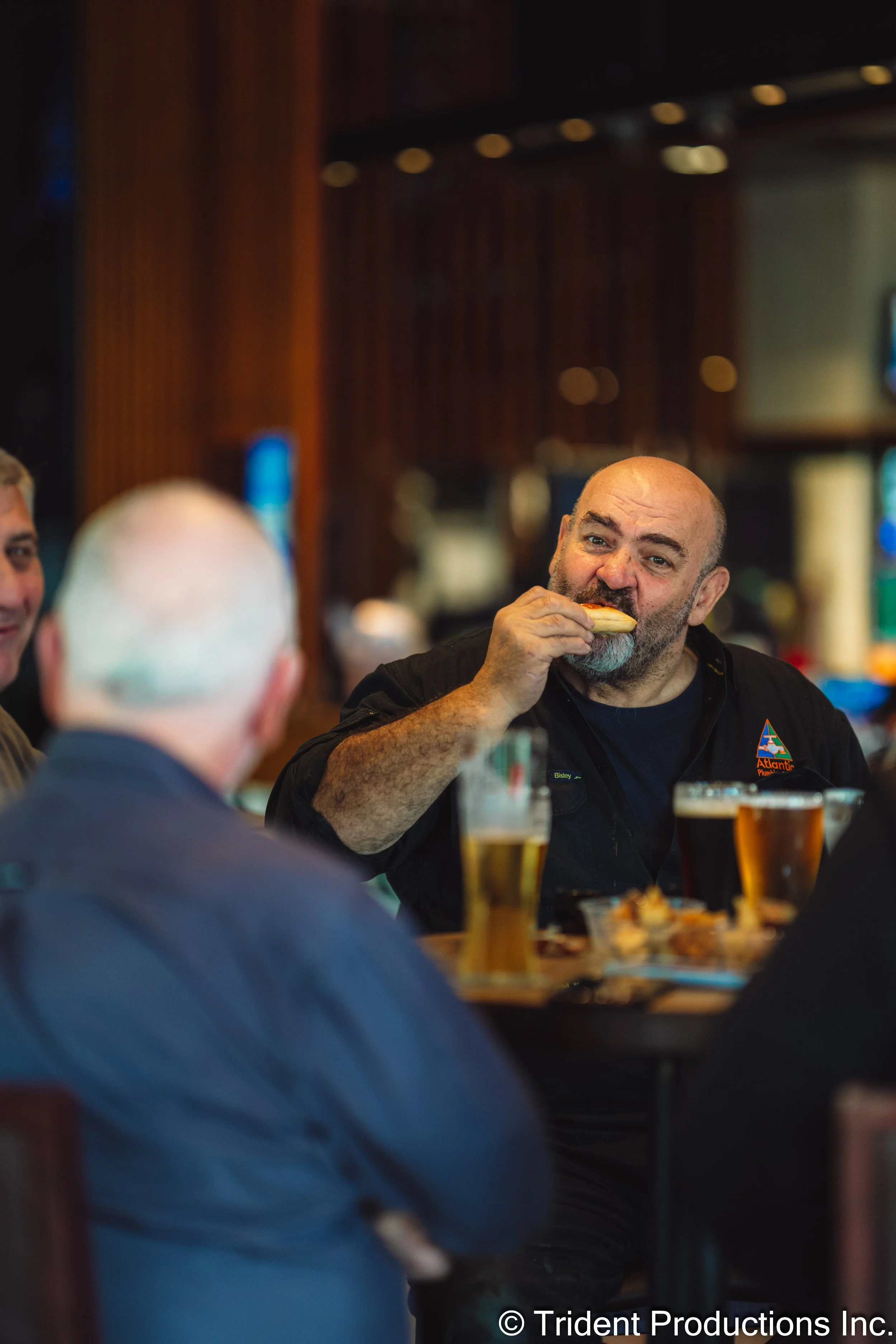 A man with a beard and bald head eating a sandwich at a bar or restaurant, with glasses of beer in front of him and other people visible in the background.