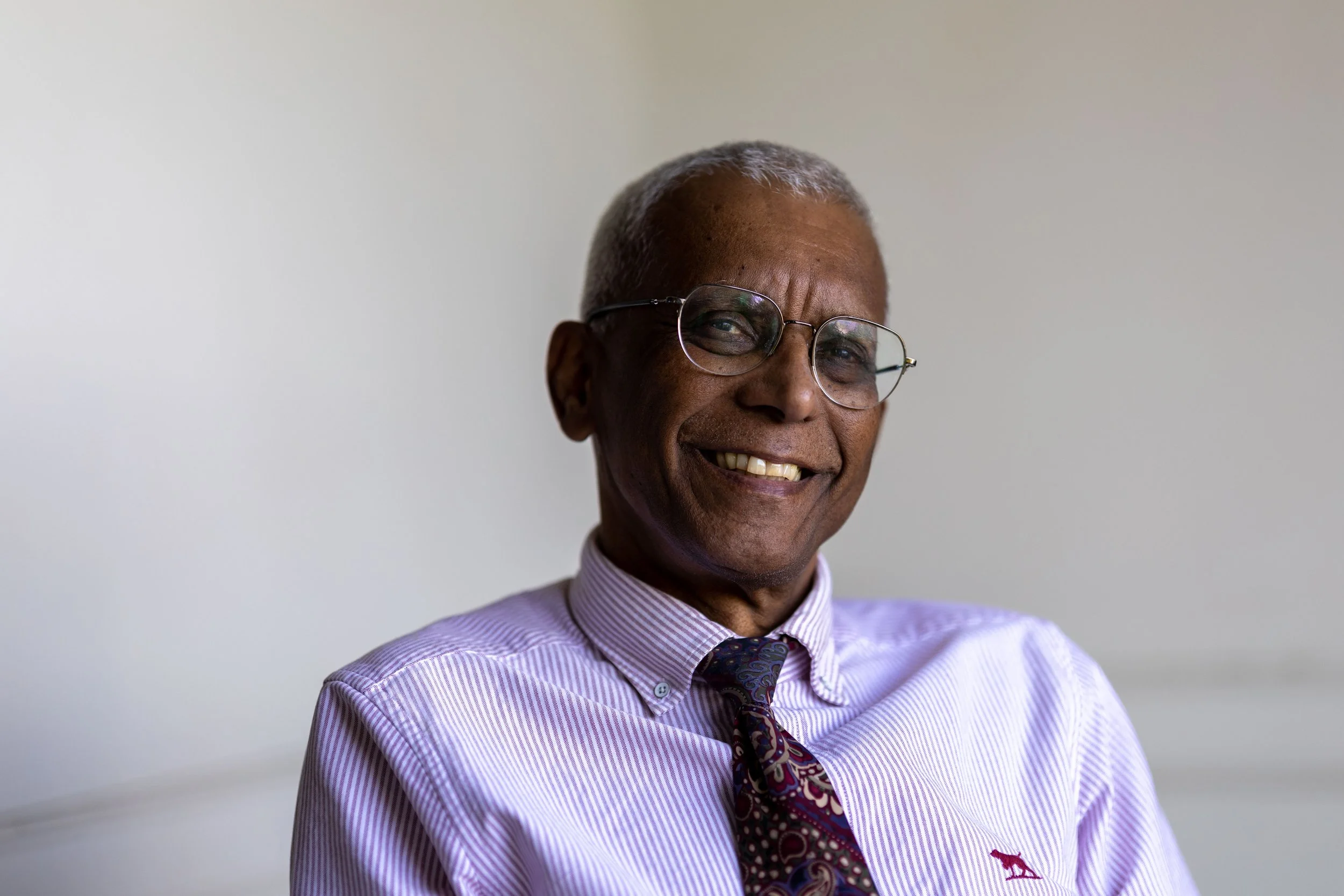 An elderly man with glasses smiling, wearing a light purple striped shirt and a patterned tie.