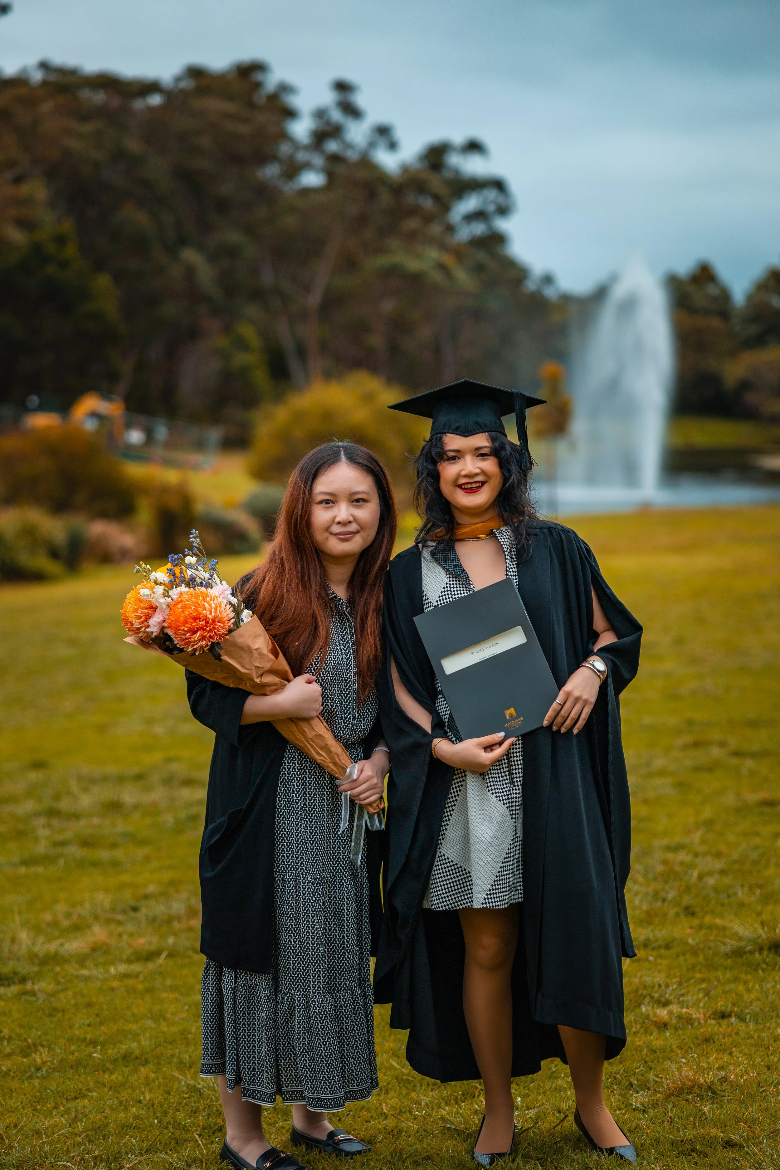 Two women, one in a graduation cap and gown, standing outdoors on a grassy field with a fountain and trees in the background, celebrating a graduation.