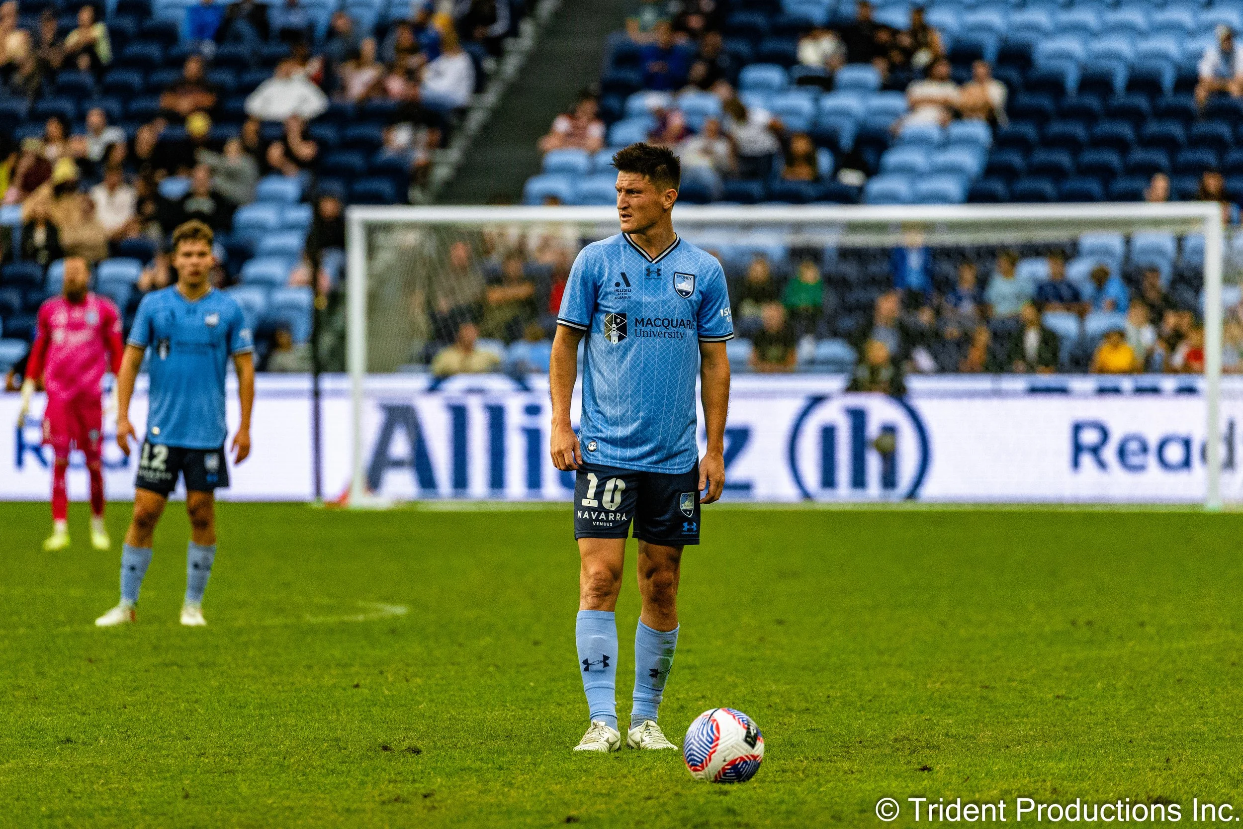 A soccer player in a light blue jersey stands on the field preparing to kick the ball, with other players and spectators in the background.