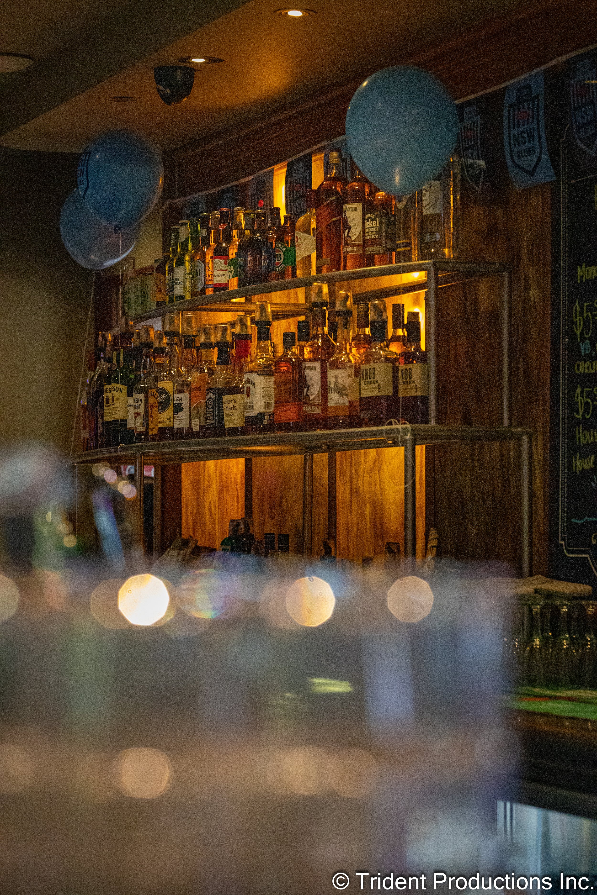 Bar shelf with various bottles of liquor, decorated with two blue balloons on each side, inside a dimly lit bar or pub.