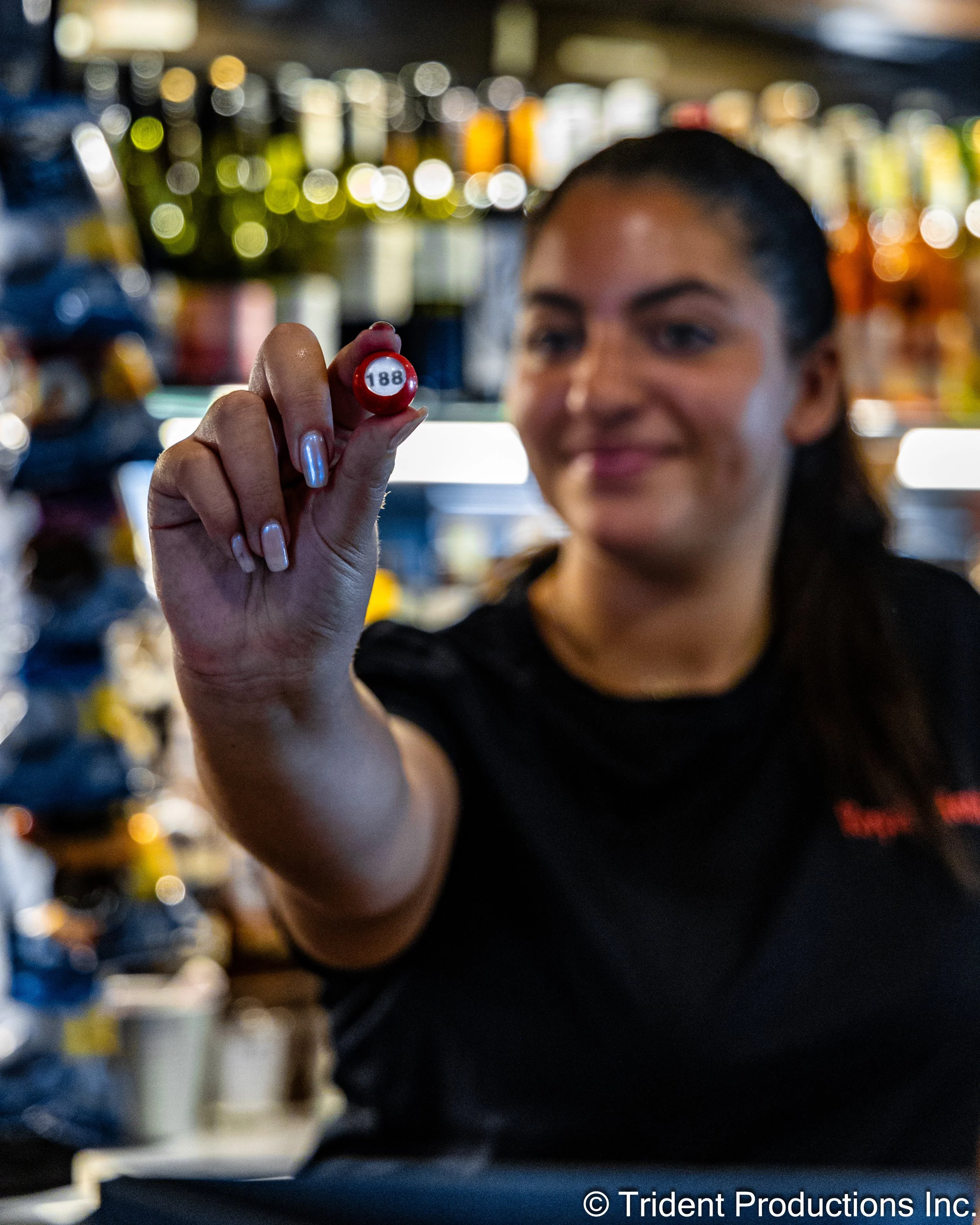 A woman in a black shirt holding a red and white billiard ball with the number 188 on it, in a bar or game room with blurred shelves of bottles in the background.